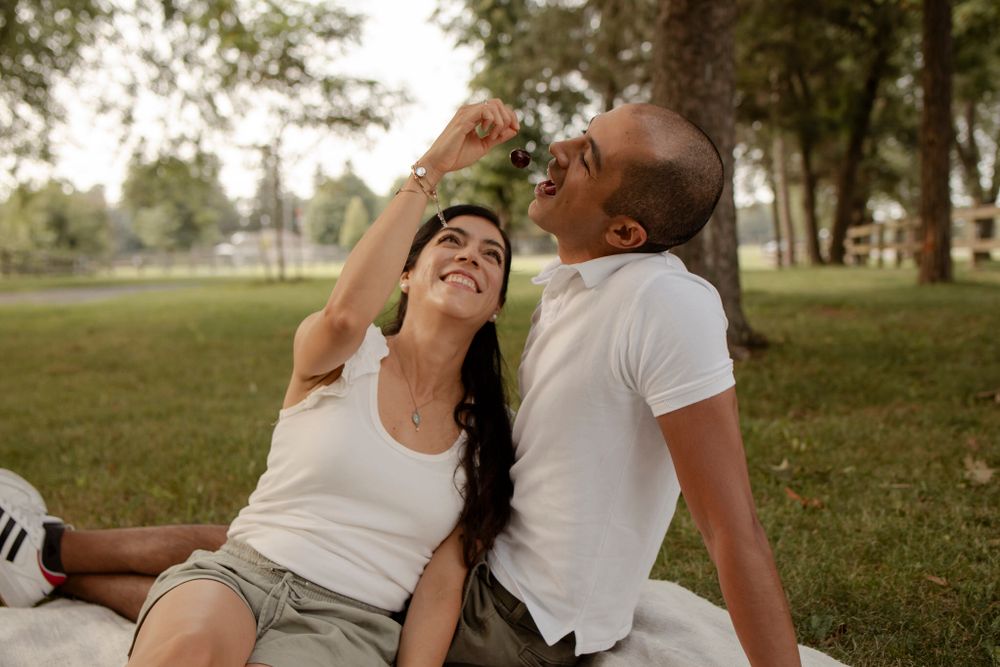 Couple sitting on a white picnic blanket in the evening sunshine, Lake Ontario Park, Kingston, ON, Ca. She feeds him a cherry playfully.