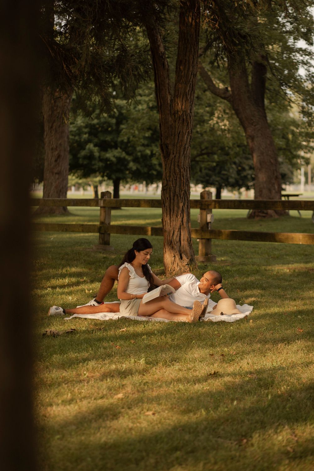 Couple sitting on a white picnic blanket in the evening sunshine, Lake Ontario Park, Kingston, ON, Ca. She reads. They smile.