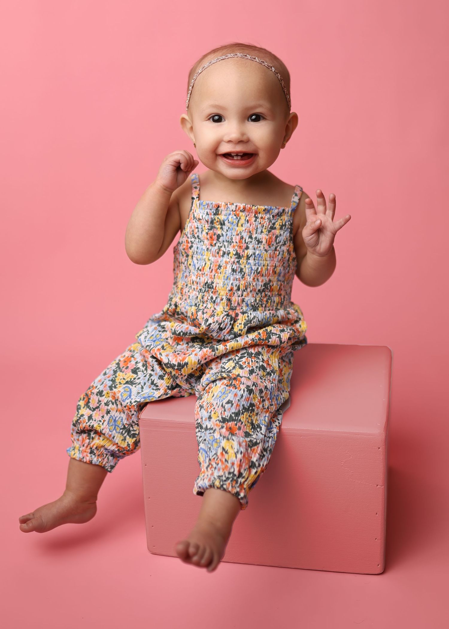 Smiling baby wearing floral overalls sits on pink cube against pink background, hands raised playfully.