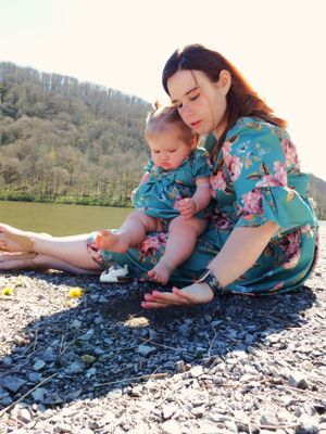 Two people in floral dresses sitting on rocky shore examining stones by the water.