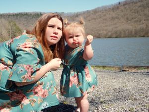 Two people in matching teal floral dresses pose together by a lake on a rocky shore.