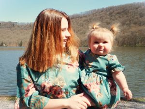 Two people in matching teal floral dresses sit by a lake with mountains in the background.