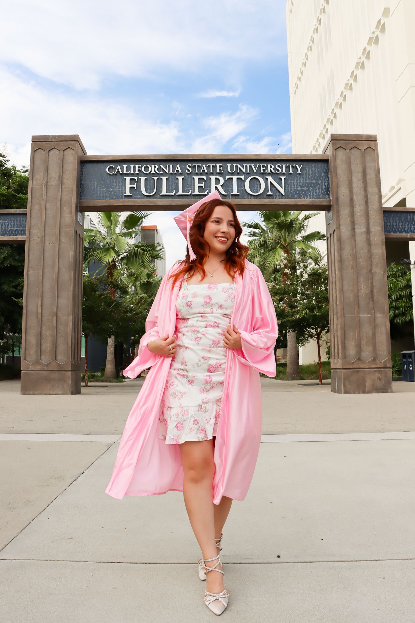 Graduate posing under Cal State Fullerton entrance sign wearing pink floral dress and robe.