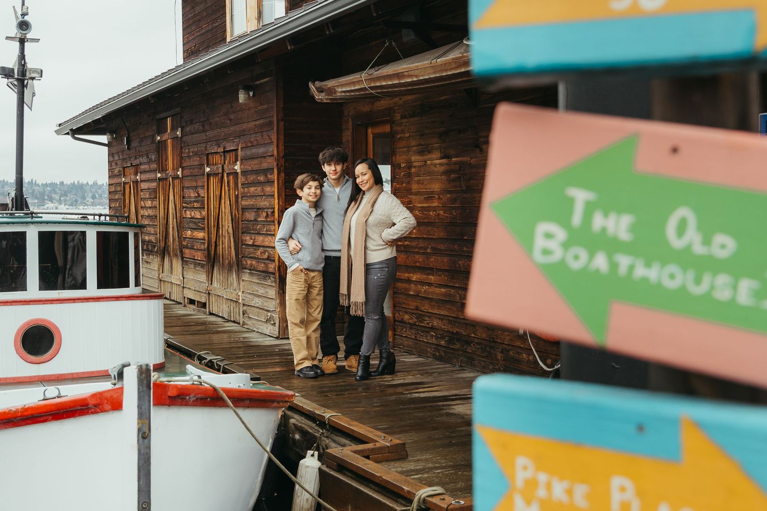 A family stands on a rustic boathouse deck with colorful signs and wooden architecture near the water.