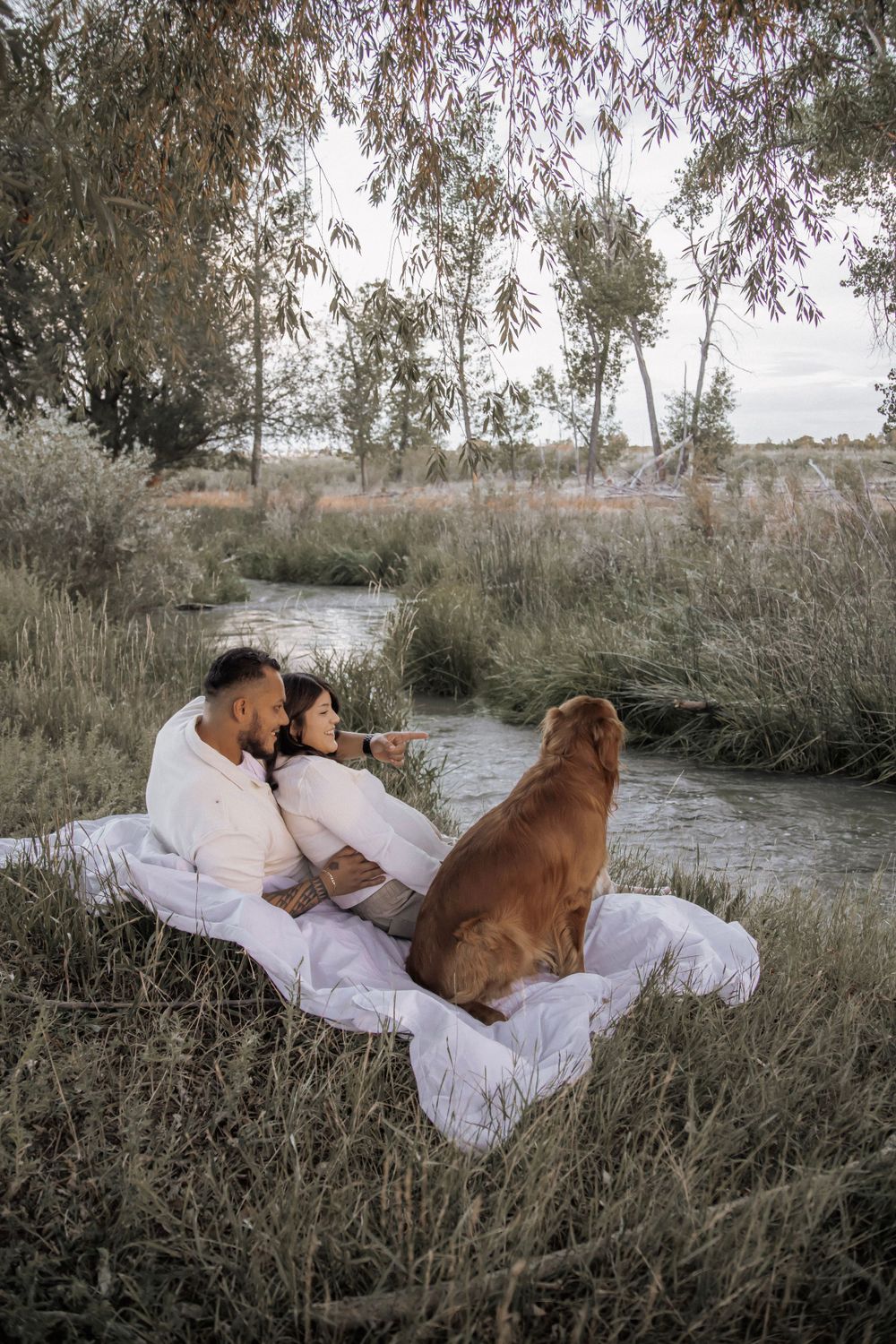 Couple snuggles with their dog under a tree by a river