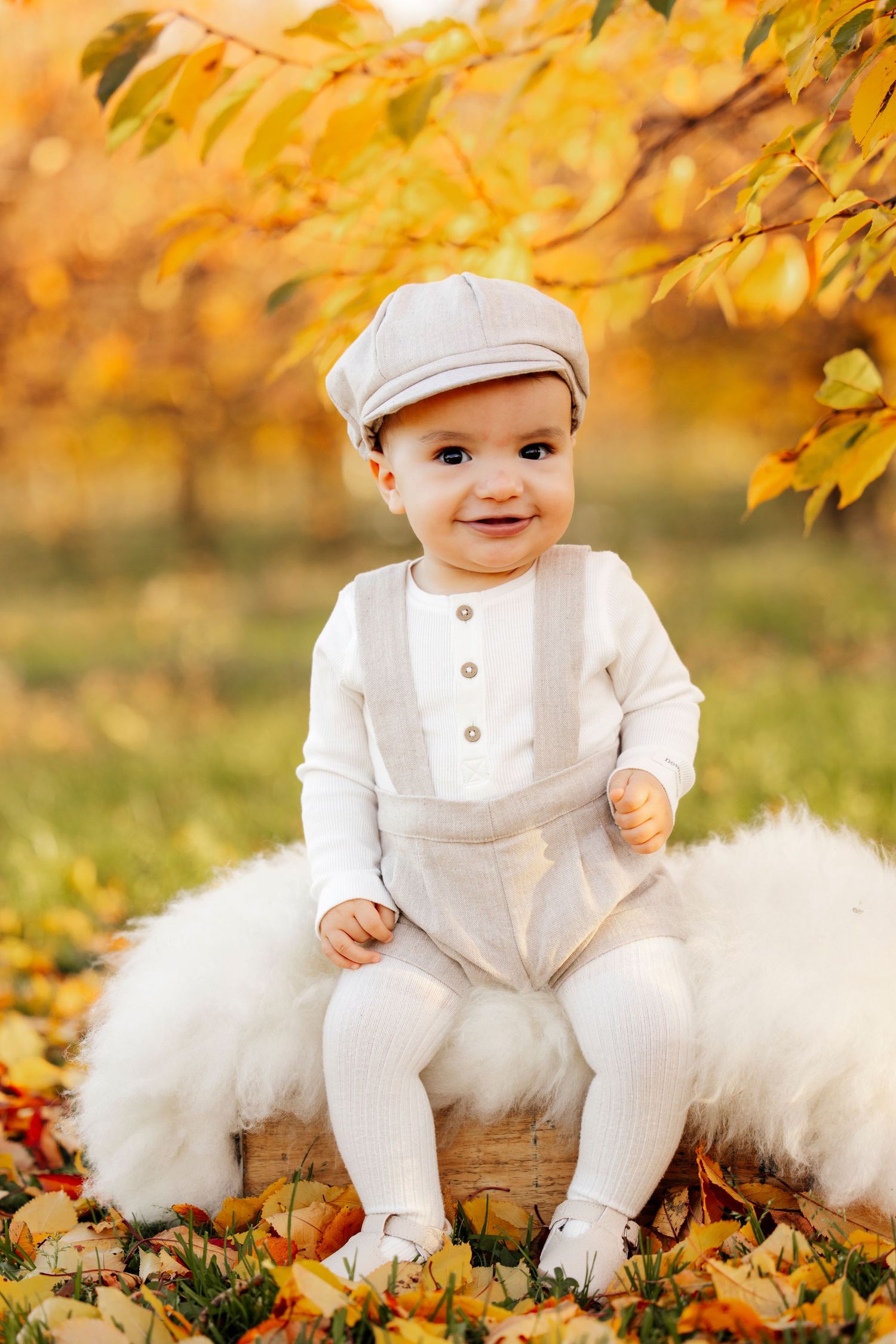 Baby in white vintage outfit and cap sitting on furry backdrop surrounded by fall leaves.
