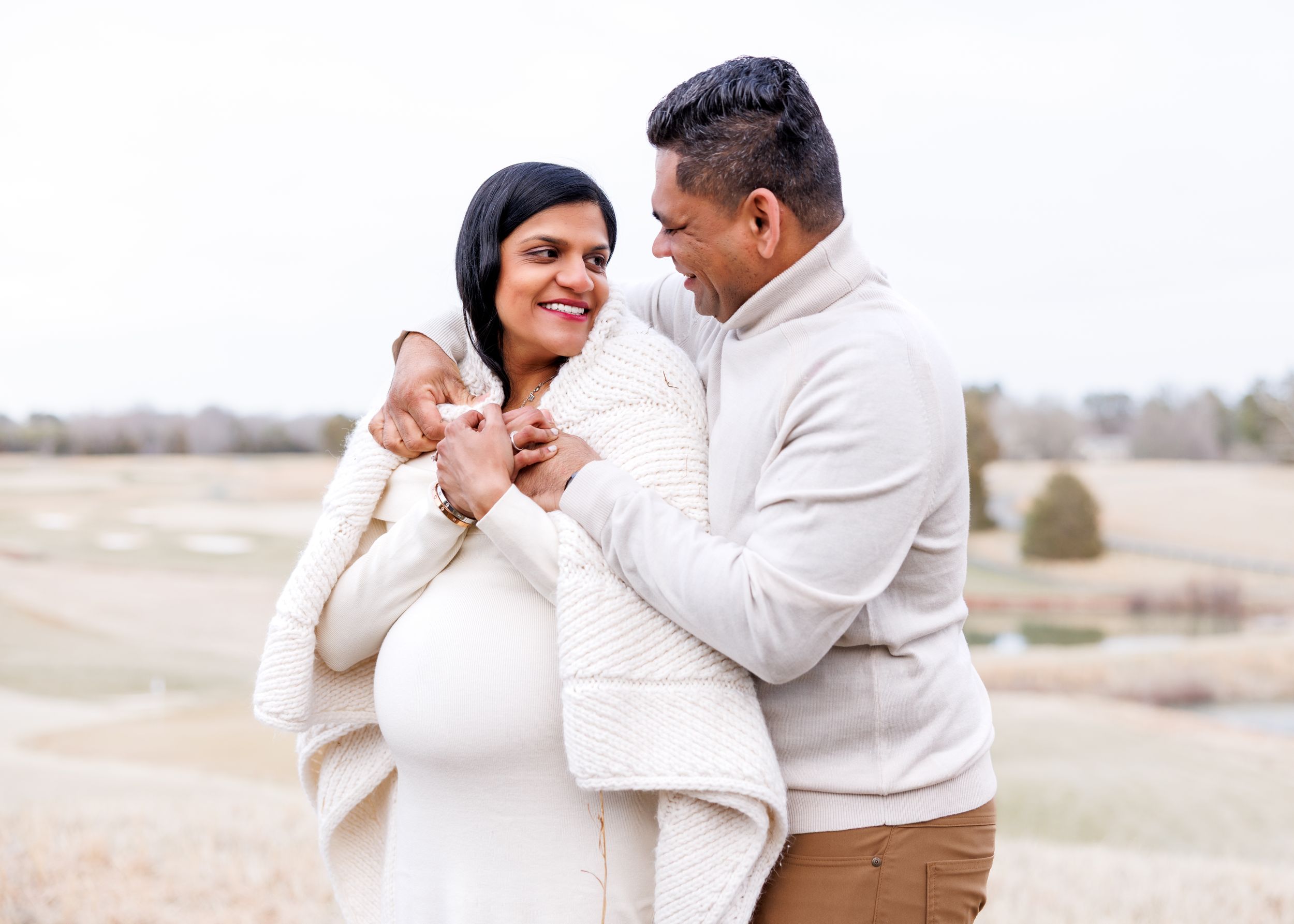 A couple wearing cream-colored sweaters embrace outdoors in a winter landscape while sharing a tender moment.