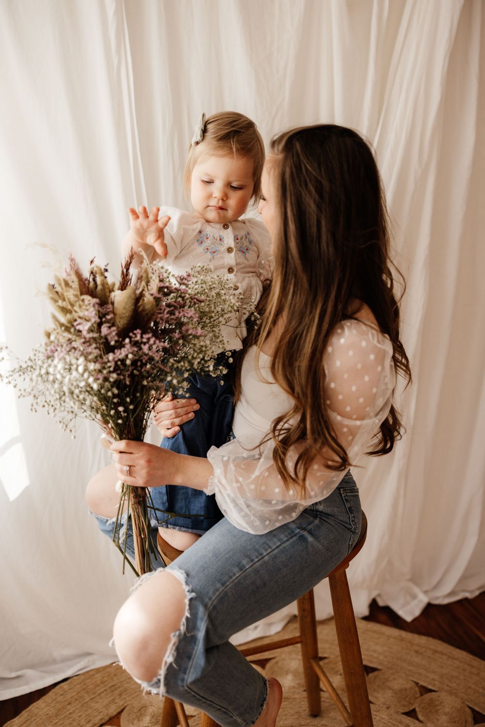 Mother and child share tender moment on wooden stool with rustic dried flower bouquet against white backdrop.