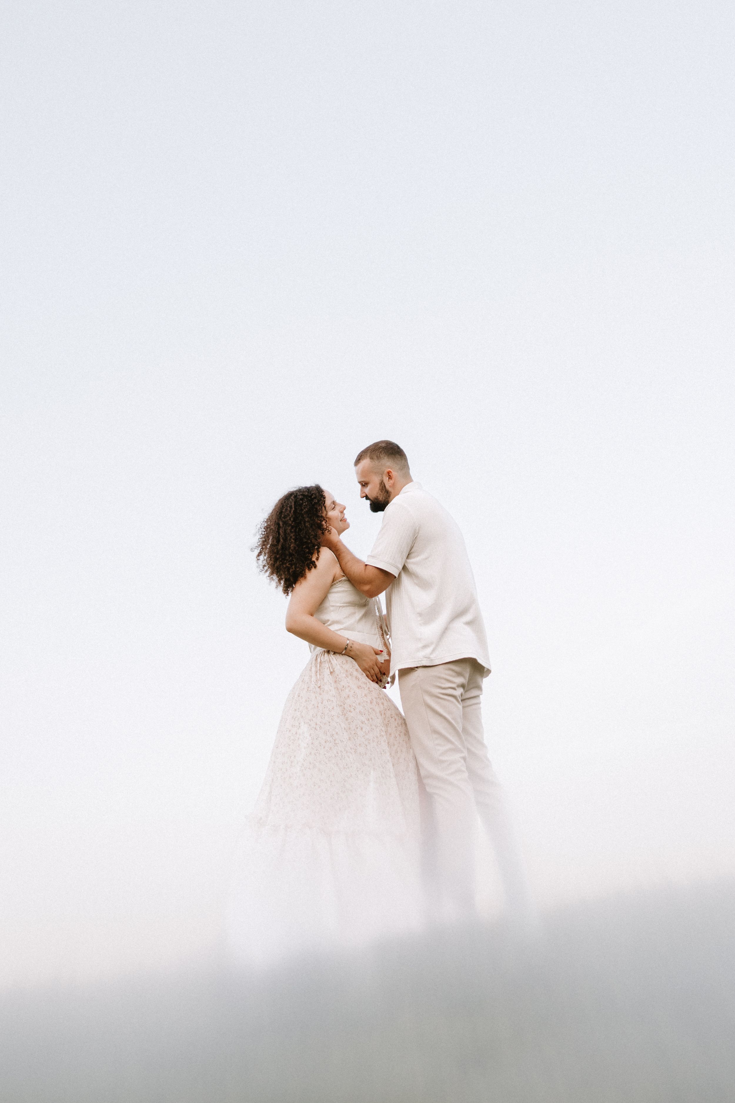 A romantic couple in white outfits embrace while posing for wedding photos along a misty mountainside.