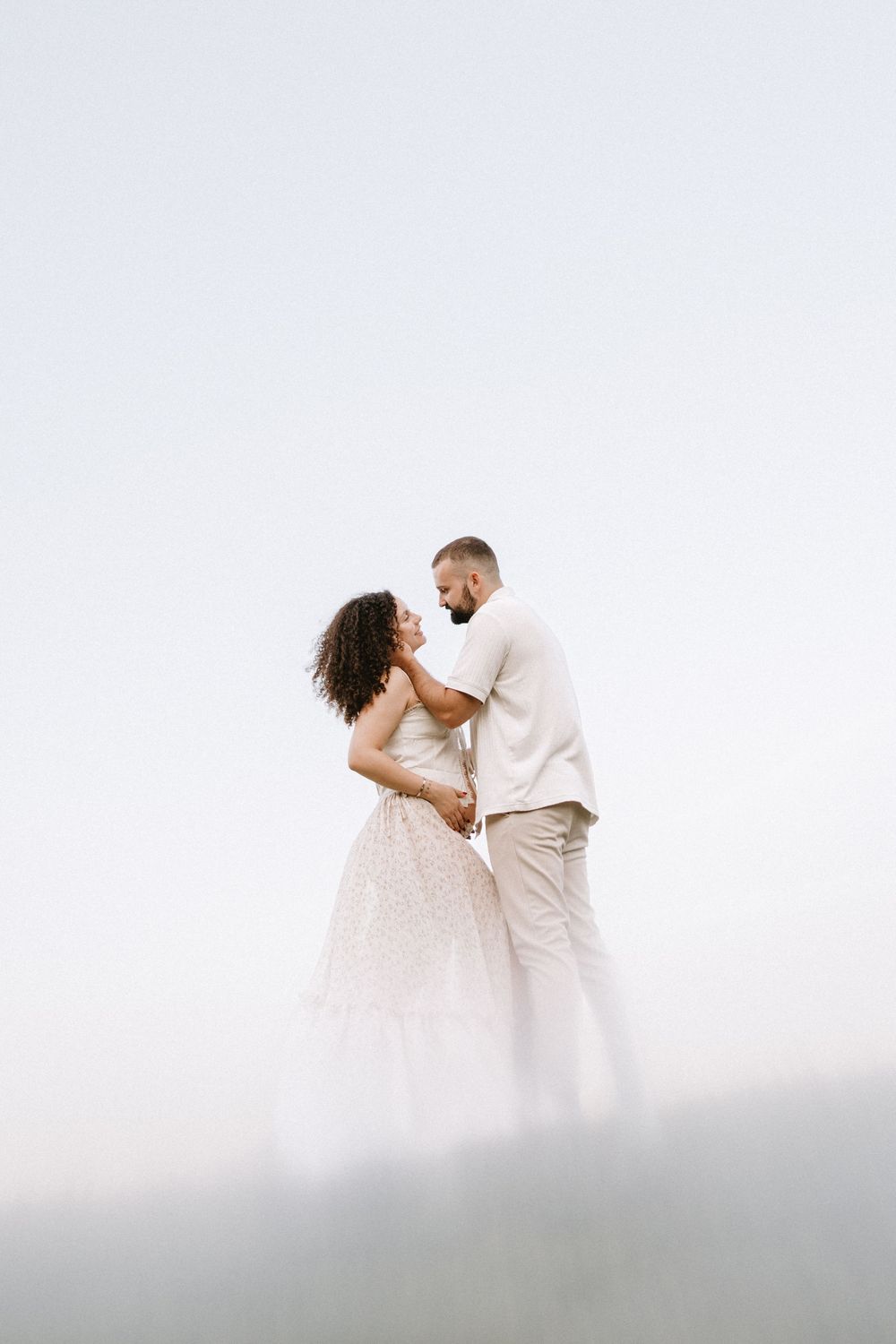 A romantic couple in white outfits embrace while posing for wedding photos along a misty mountainside.