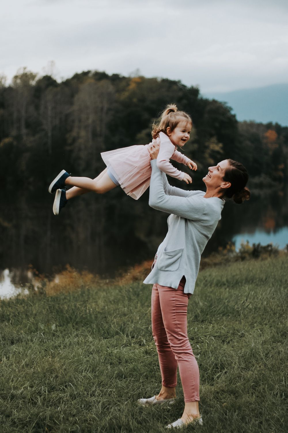Joyful outdoor portrait of parent lifting child in pink dress by lakeside during autumn evening.
