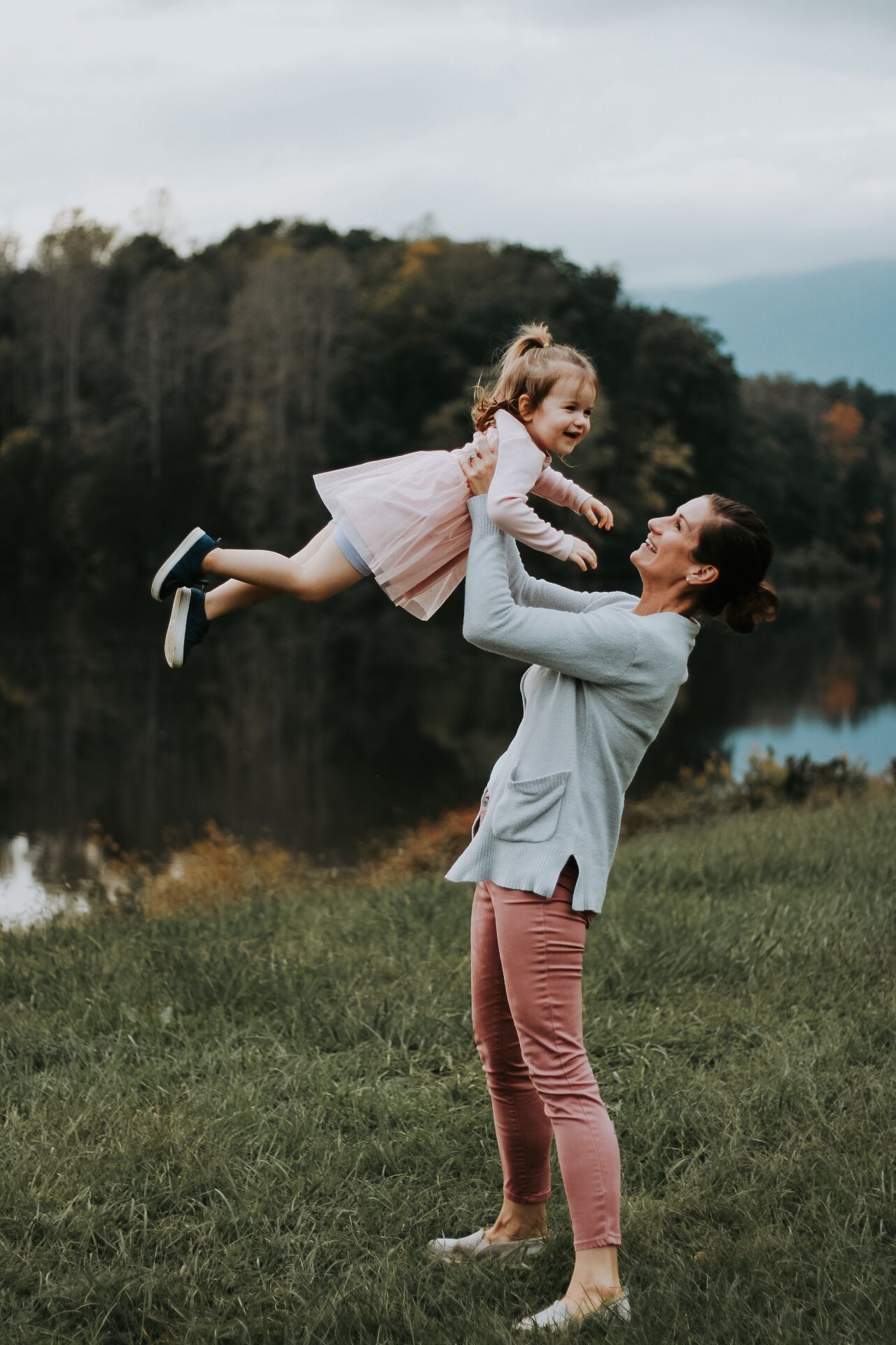 Joyful outdoor portrait of parent lifting child in pink dress by lakeside during autumn evening.