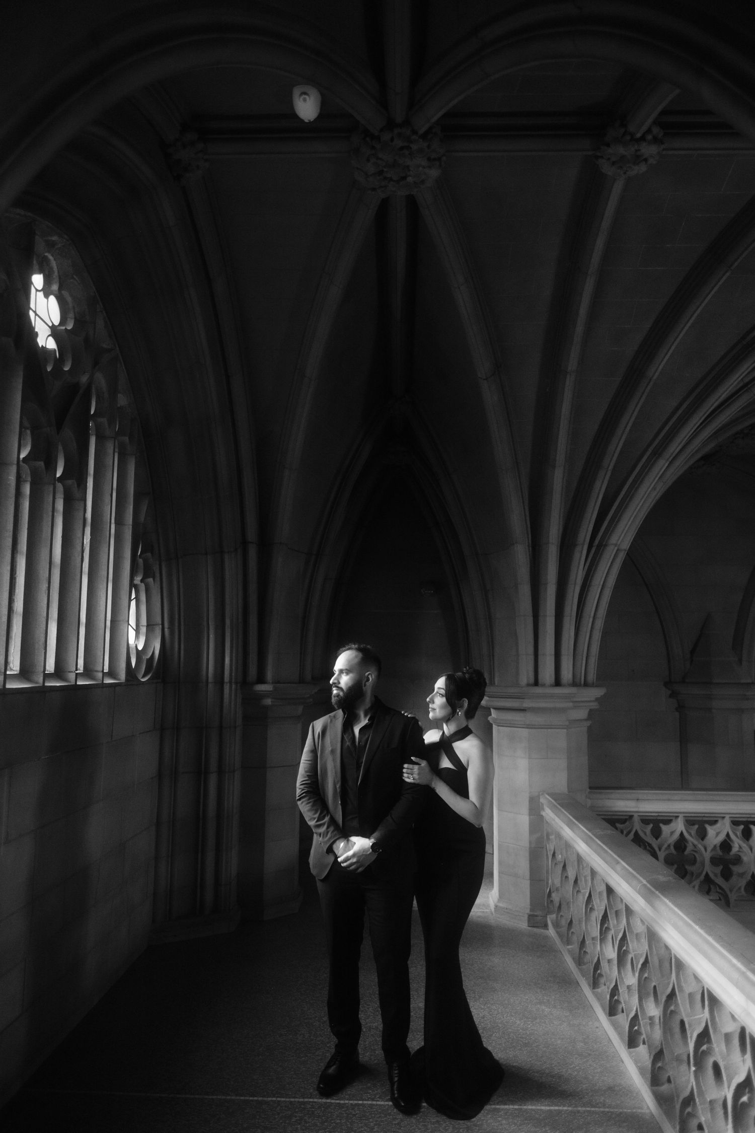 A couple poses elegantly in a gothic archway corridor with dramatic lighting and architectural details.