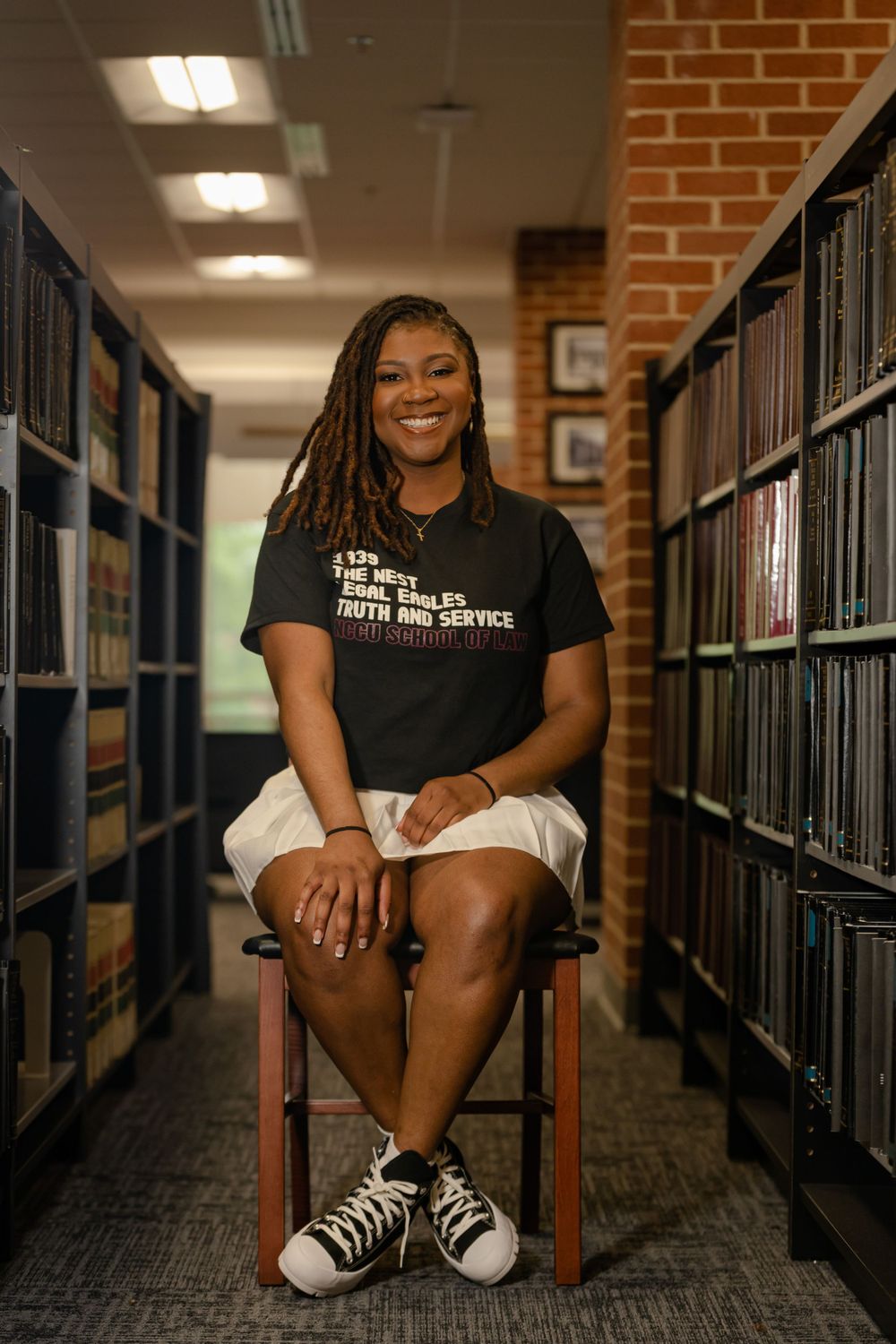 A cheerful person sits on a wooden chair in a library aisle surrounded by bookshelves and wearing a dark t-shirt.