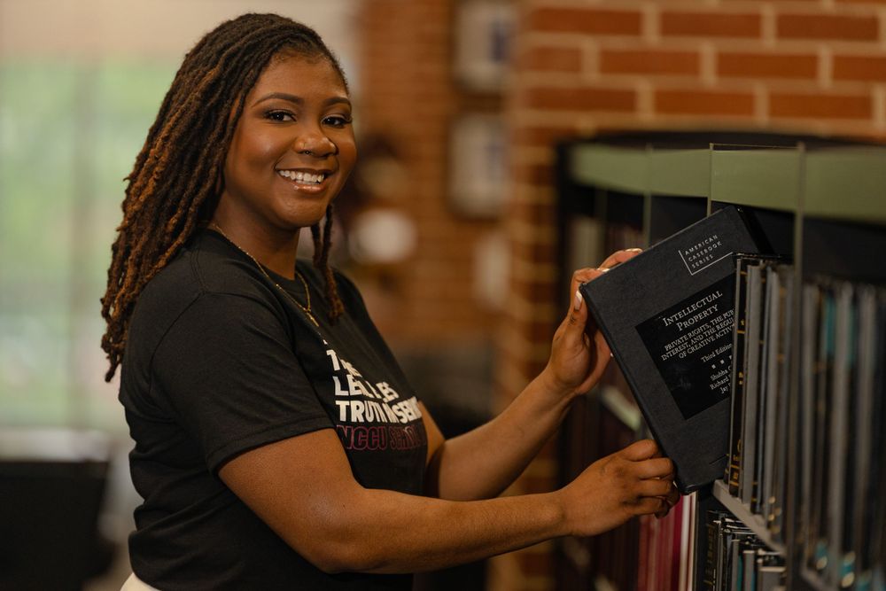 A smiling employee in a black t-shirt organizes books on a shelf at a store.