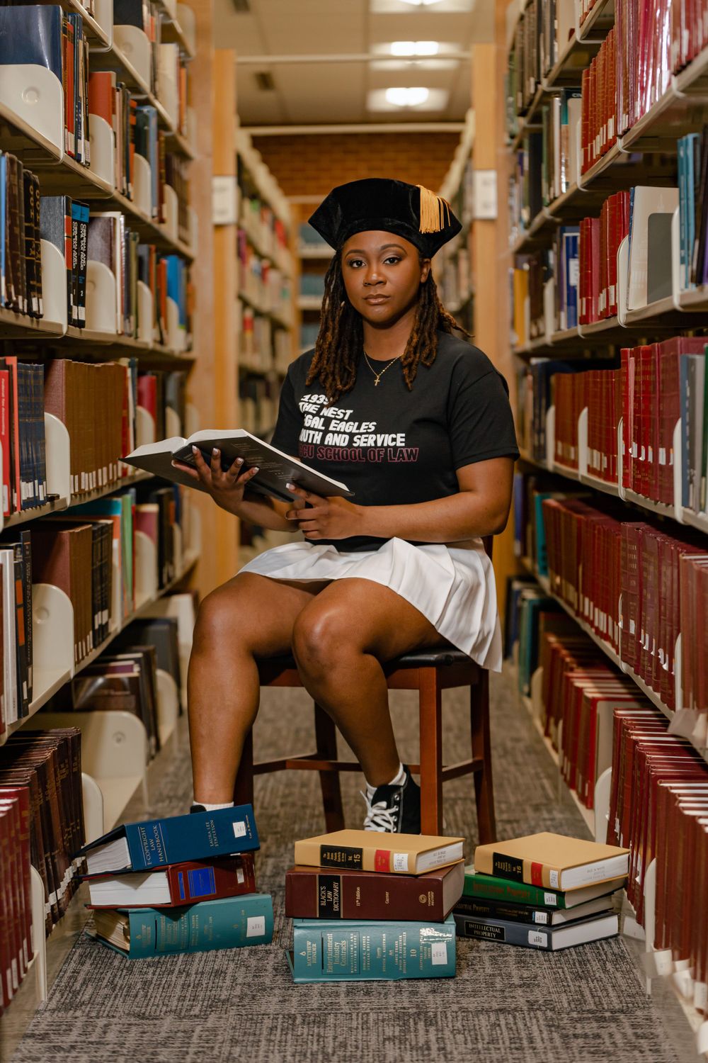 Graduate student in academic regalia studies in library stacks with books stacked at feet while seated on wooden stool.