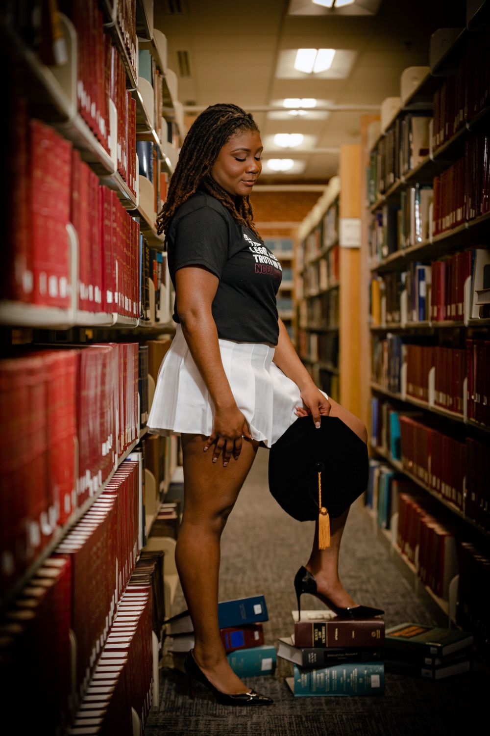 Student poses between library bookshelves wearing black t-shirt and white skirt while holding graduation cap.