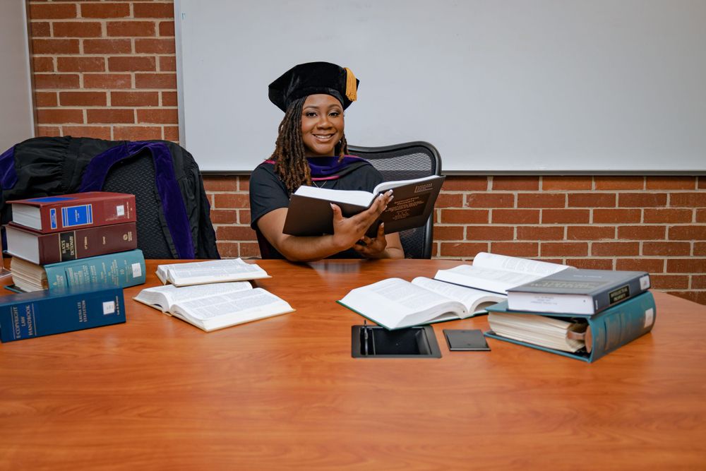 A student wearing a graduation cap and gown studies at a wooden table surrounded by stacks of textbooks and binders.