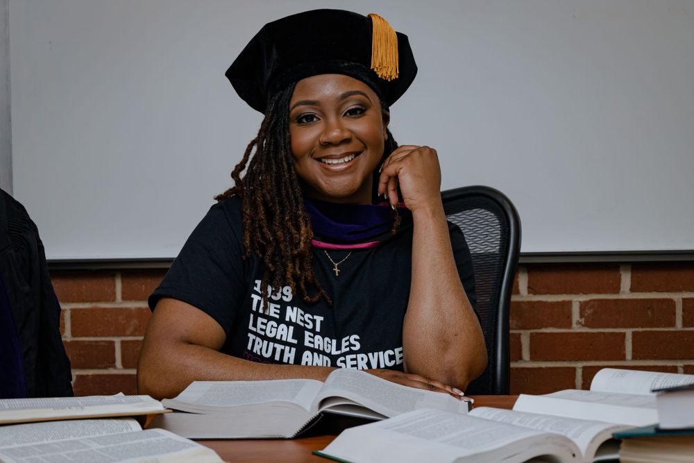 Student wearing graduation cap sits at desk with books while resting head on hand and smiling.