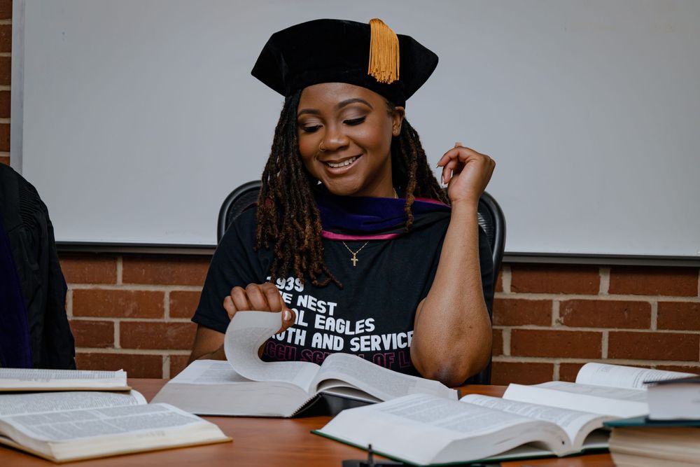 A student in a graduation cap sits at a desk with textbooks, smiling while studying at a brick-walled classroom.