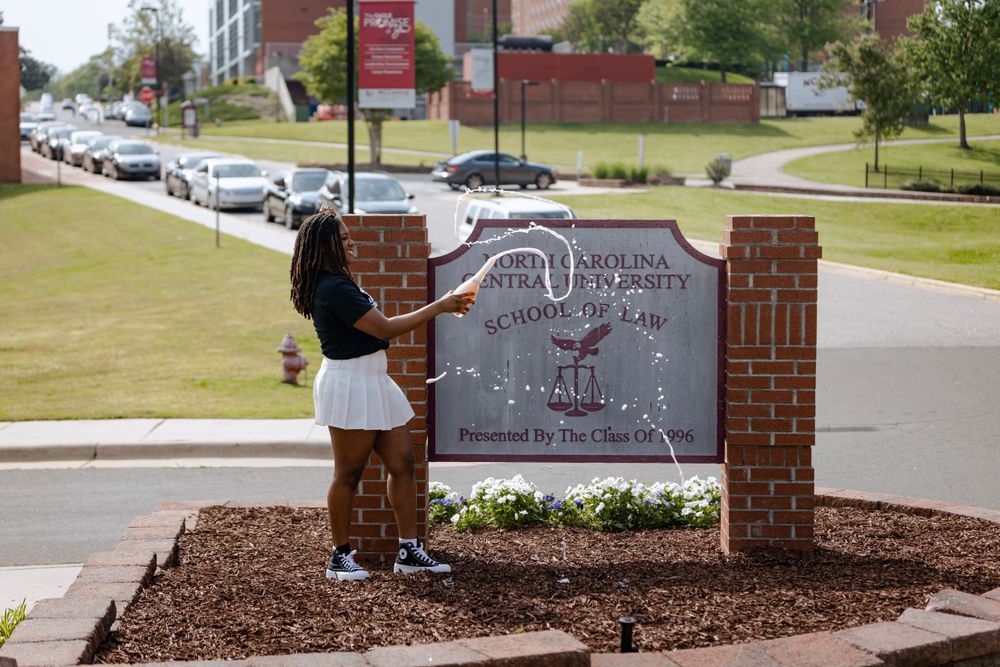 Student in white skirt stands by brick entrance sign at a university campus with mulch landscaping and parked cars.