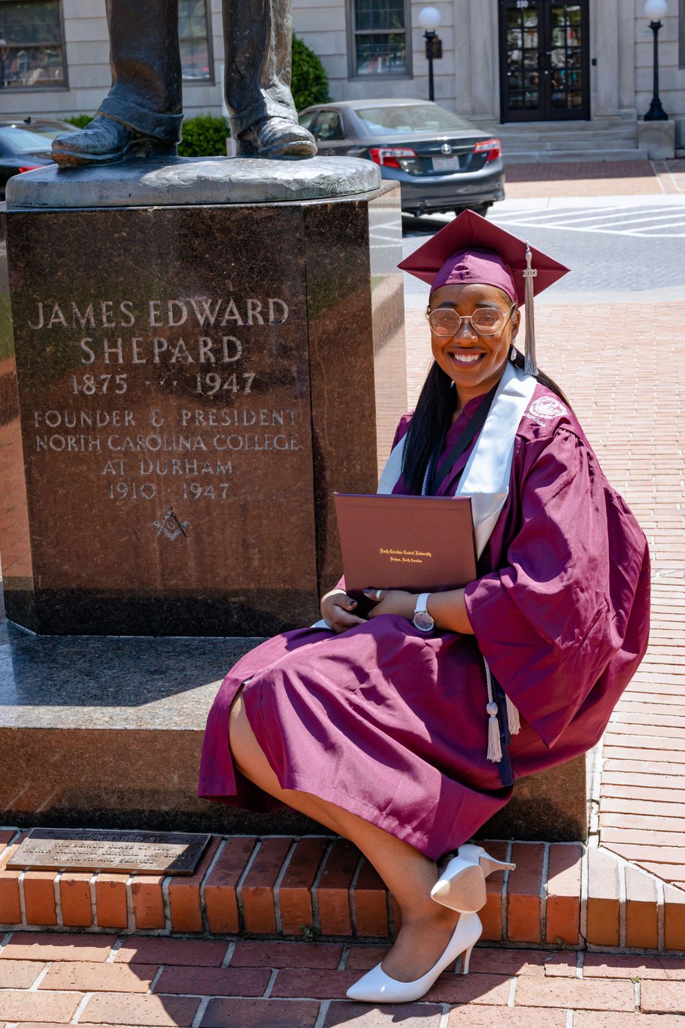 Graduate poses with diploma by James Edward Shepard statue in maroon academic regalia at North Carolina Central University.
