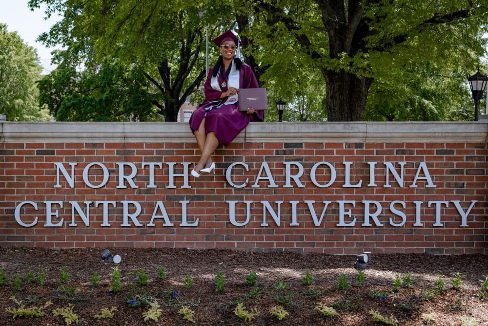 A graduate wearing purple and white regalia sits on the brick North Carolina Central University entrance sign.