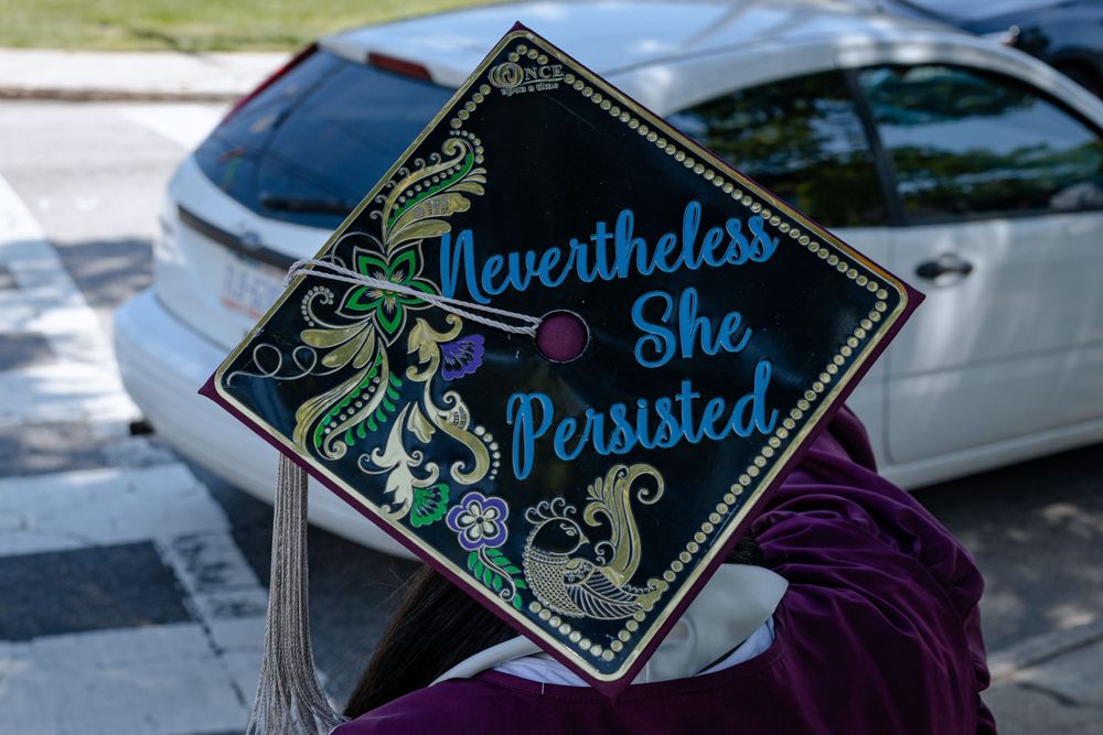 A decorated graduation cap with "Nevertheless She Persisted" written in blue glitter letters and floral designs.