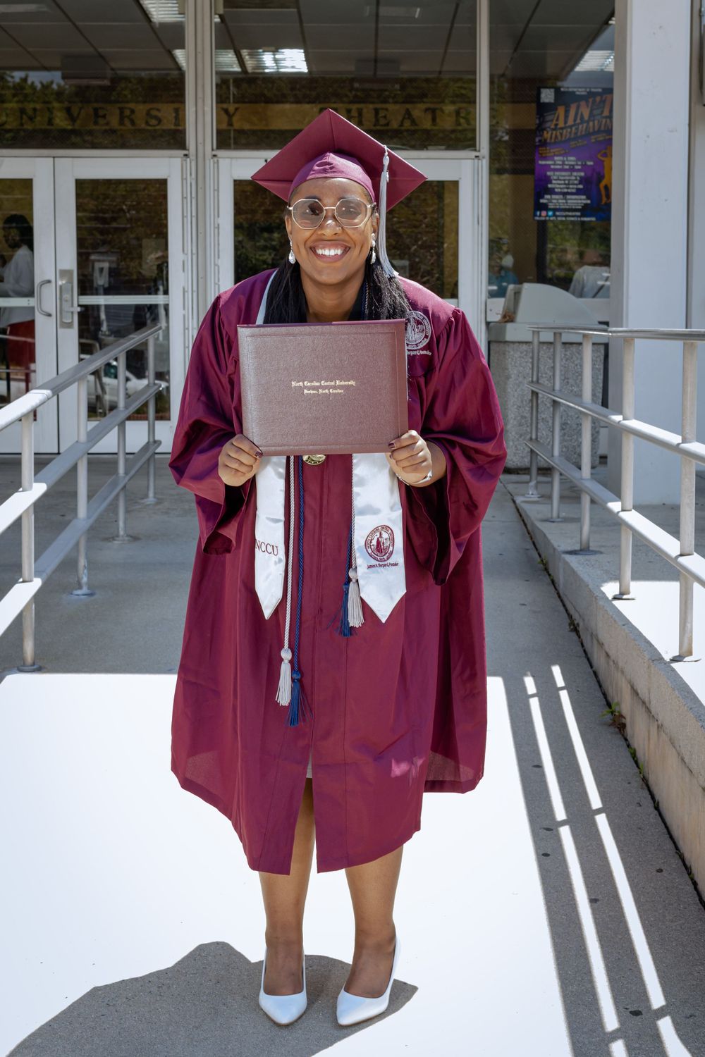 Graduate in maroon cap and gown holds diploma while standing on walkway outside building entrance.
