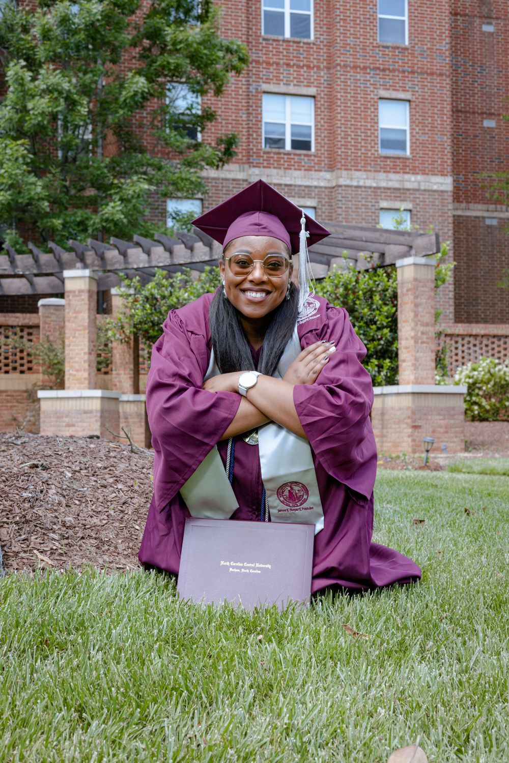 A graduate in maroon cap and gown sits on grass with their diploma on a college campus with brick buildings behind.