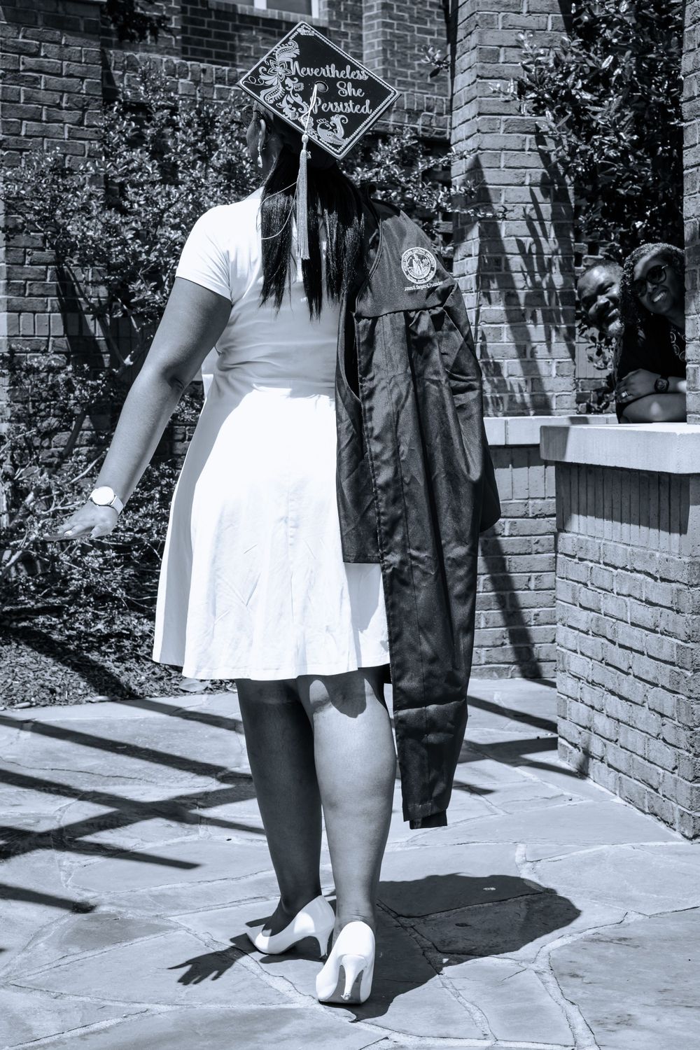 Black and white graduation photo showing graduate in white dress and decorated cap with gown draped nearby.