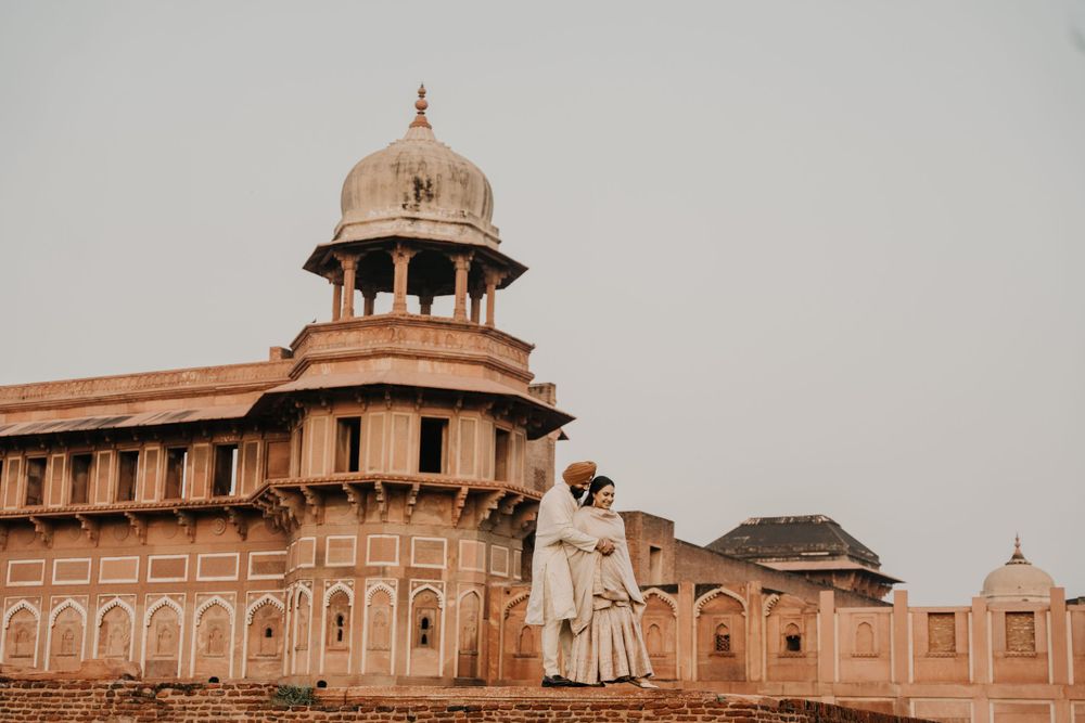Couple in traditional attire stands at historic Agra Fort in India with ornate domed architecture in warm sunset light.