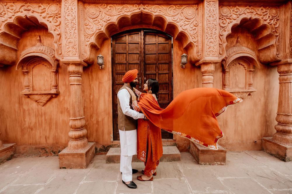 A couple in traditional Indian attire stands by an ornate terracotta doorway as orange fabric flows in the wind.
