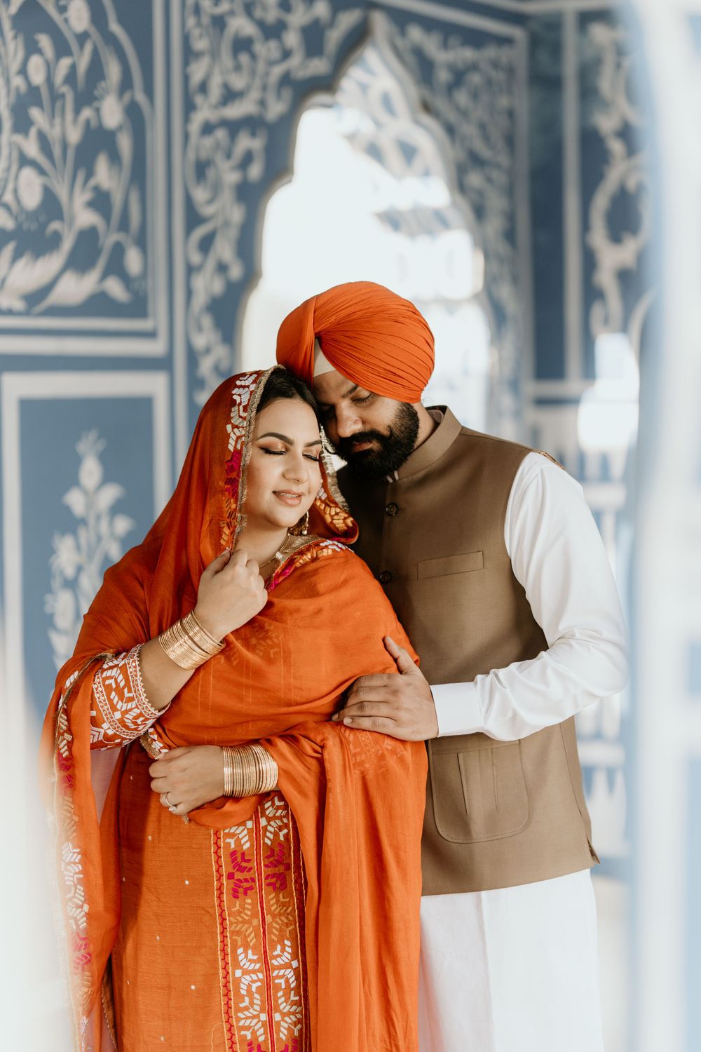 A couple in traditional Sikh wedding attire embracing in front of an ornately decorated blue and white wall.
