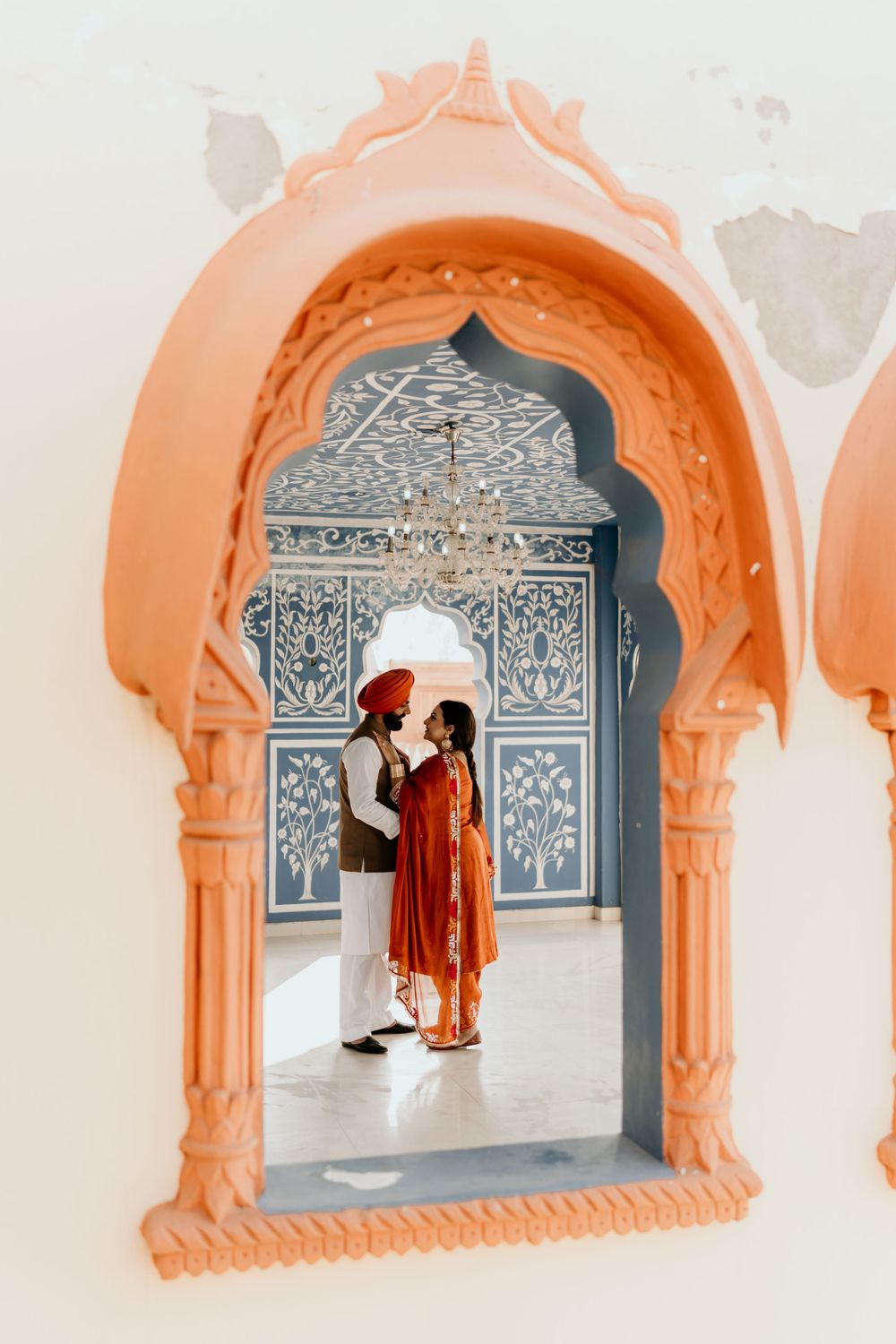 Two people in traditional Indian attire stand beneath an ornate archway with peach-colored frame and intricate silver details.
