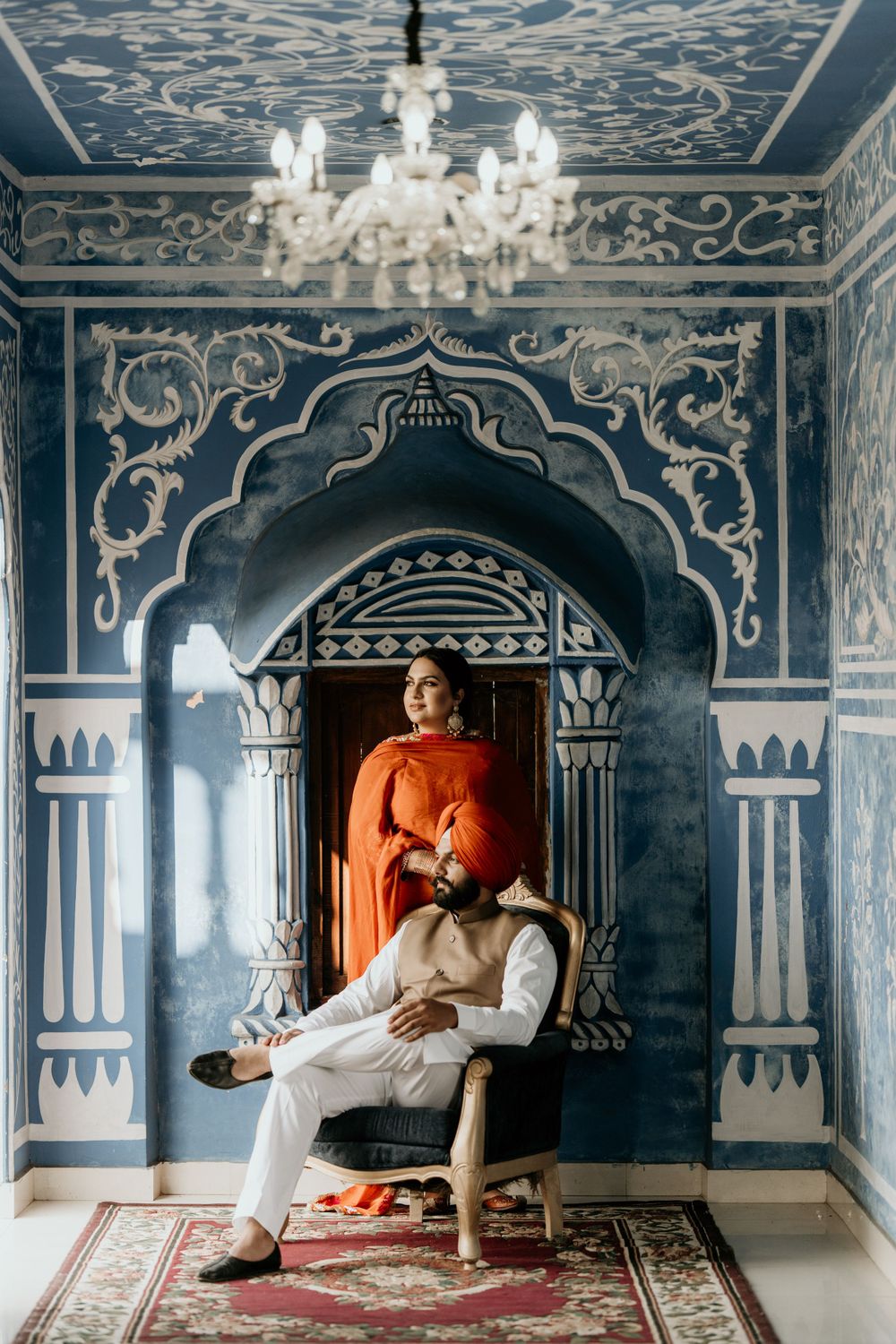 Elegant interior with ornate blue walls, crystal chandelier and decorative archway framing two people in white and orange attire.