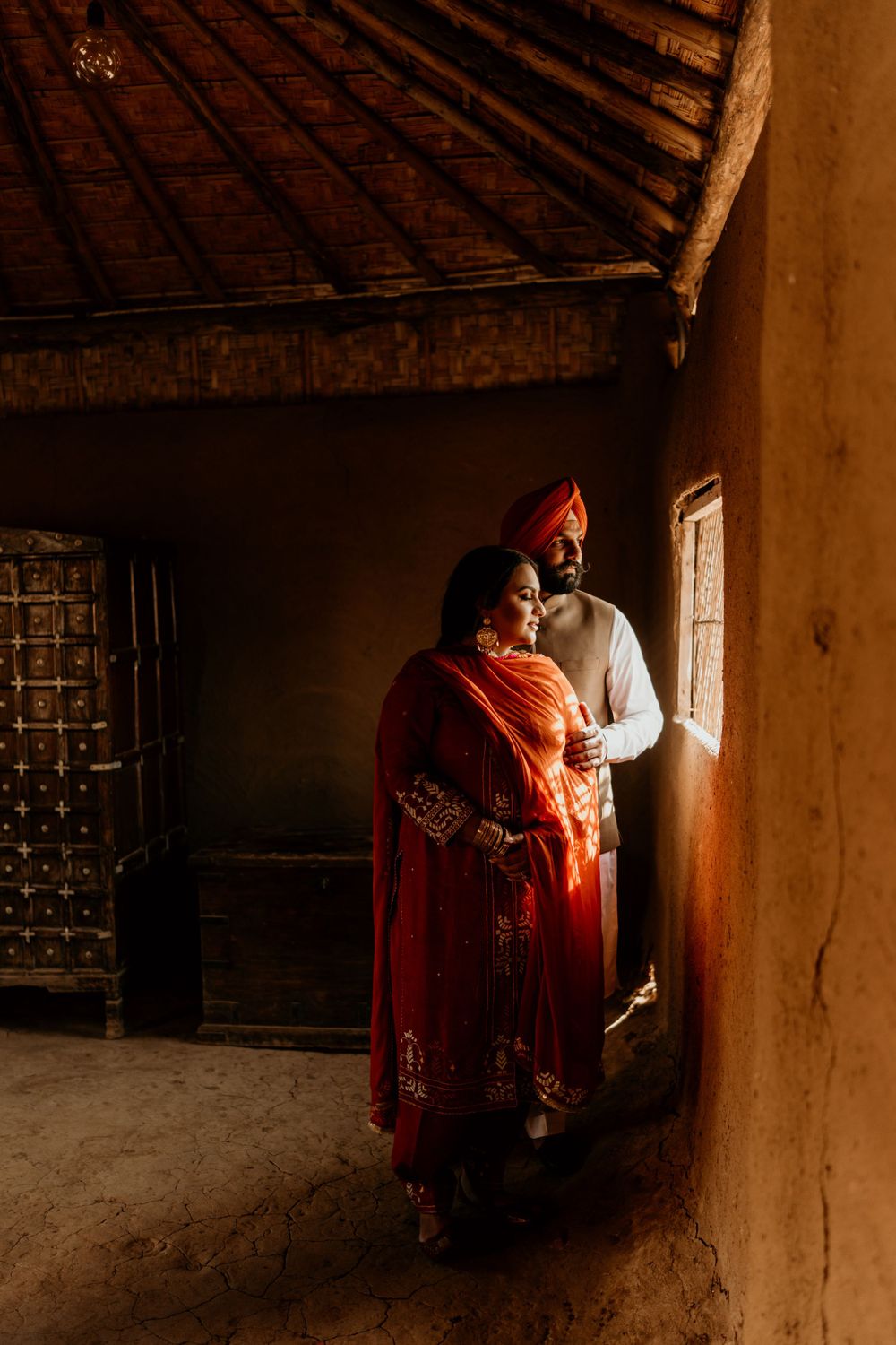 Traditional Indian couple stands in warm sunlight by window in rustic home interior with wooden beams.