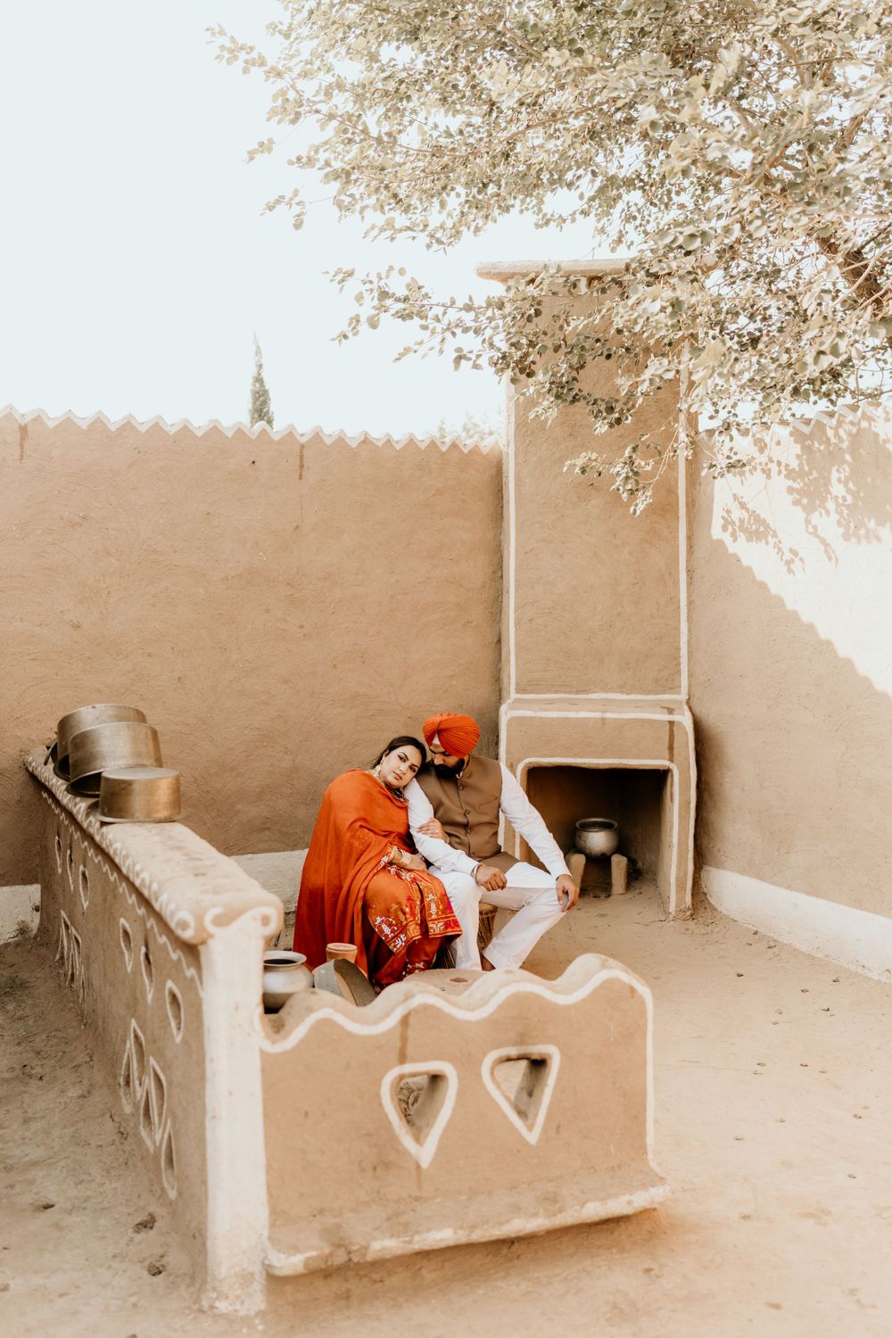 People rest on a traditional adobe bench with heart designs under a tree beside an earthen wall.