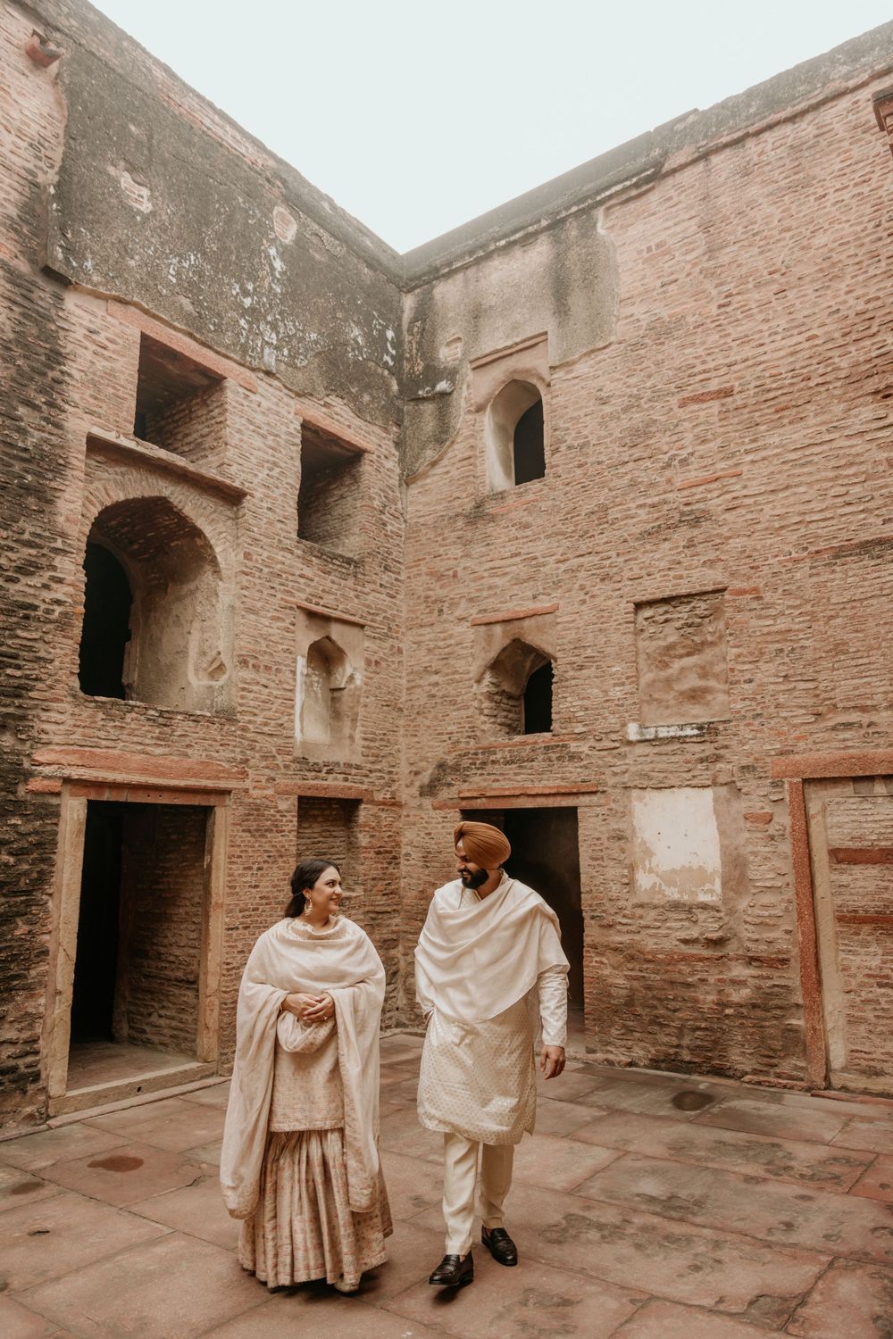 Two people in traditional Indian attire standing inside an ancient brick courtyard with arched doorways and windows.
