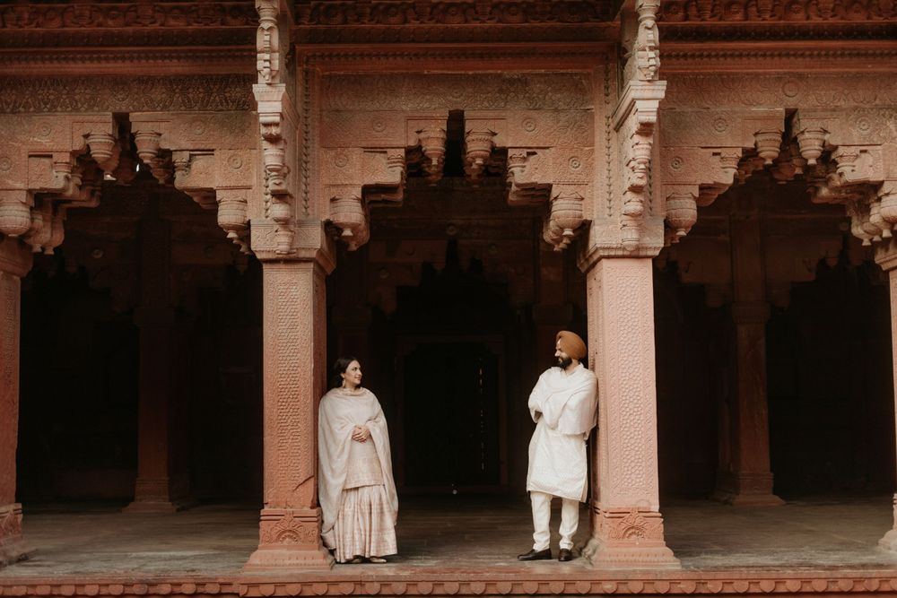 Two figures in traditional attire stand beneath ornate carved stone arches in an ancient Indian palace courtyard.