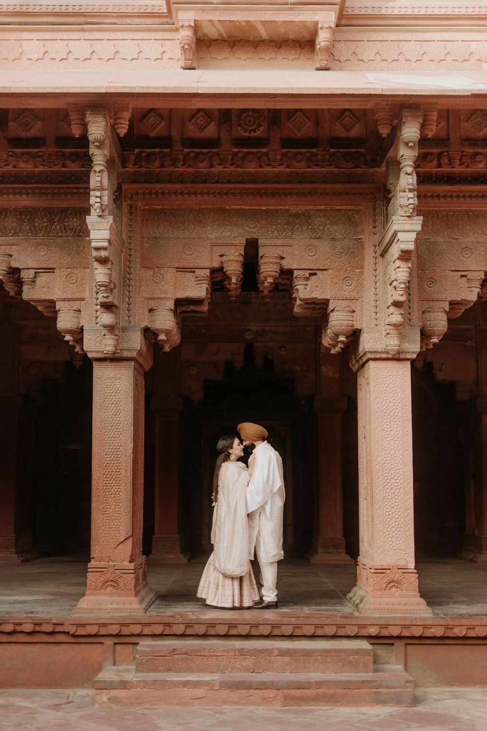 Two people in traditional clothing stand beneath ornate carved pillars and arches of an ancient Indian palace courtyard.