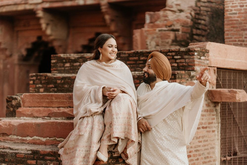 Two people wearing traditional South Asian shawls share a candid moment while sitting on brick architecture steps.
