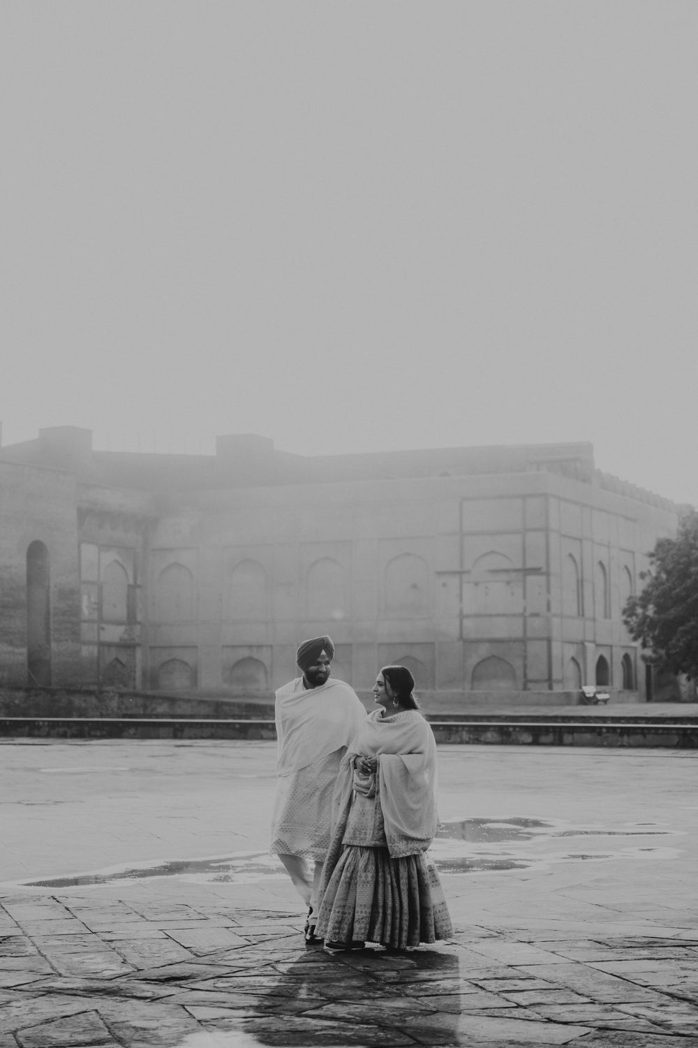Two figures wrapped in shawls stand together in a misty courtyard with historic architecture in the background.