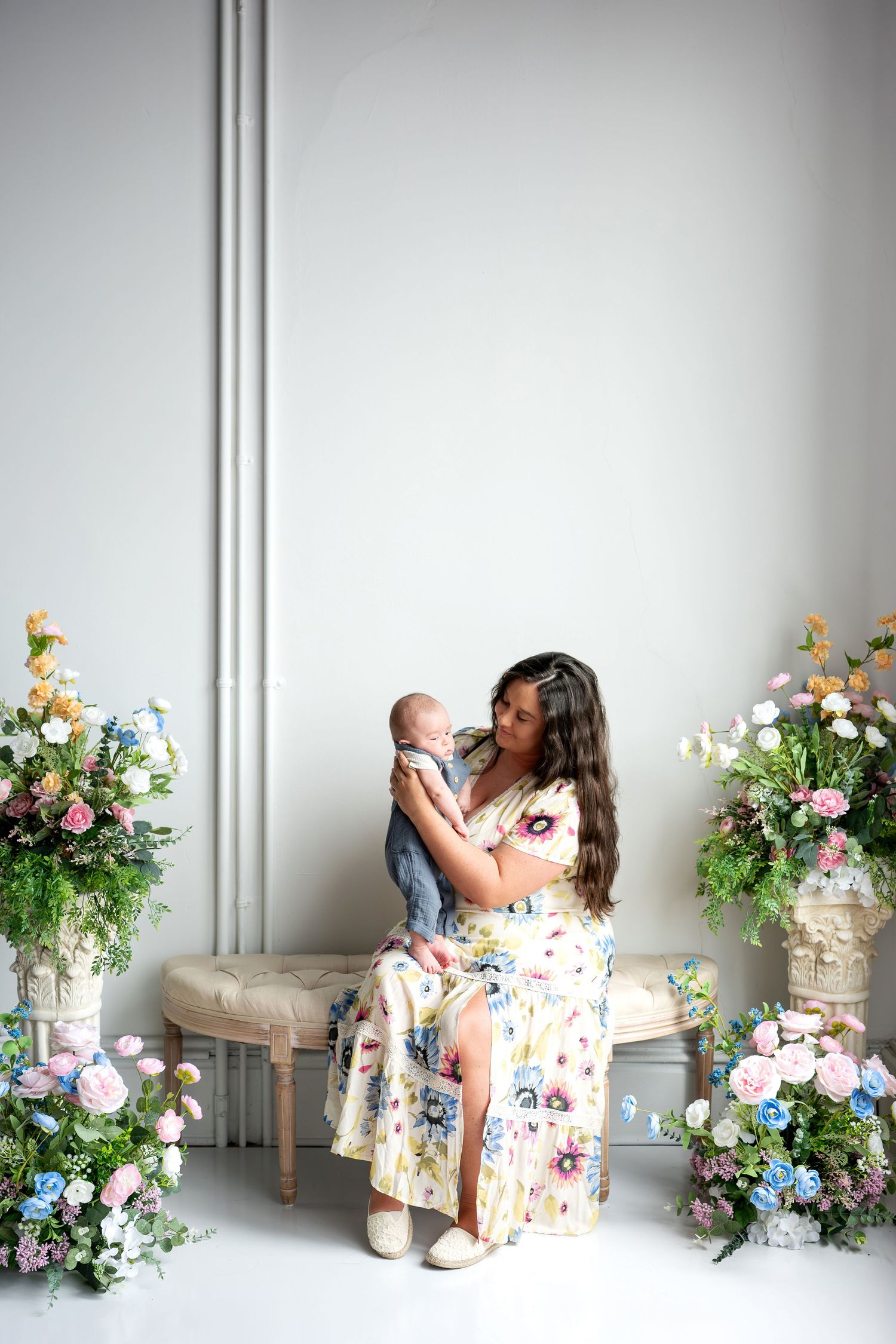 A mother in a floral dress sits on a bench holding her newborn baby, surrounded by colorful flower arrangements indoors.