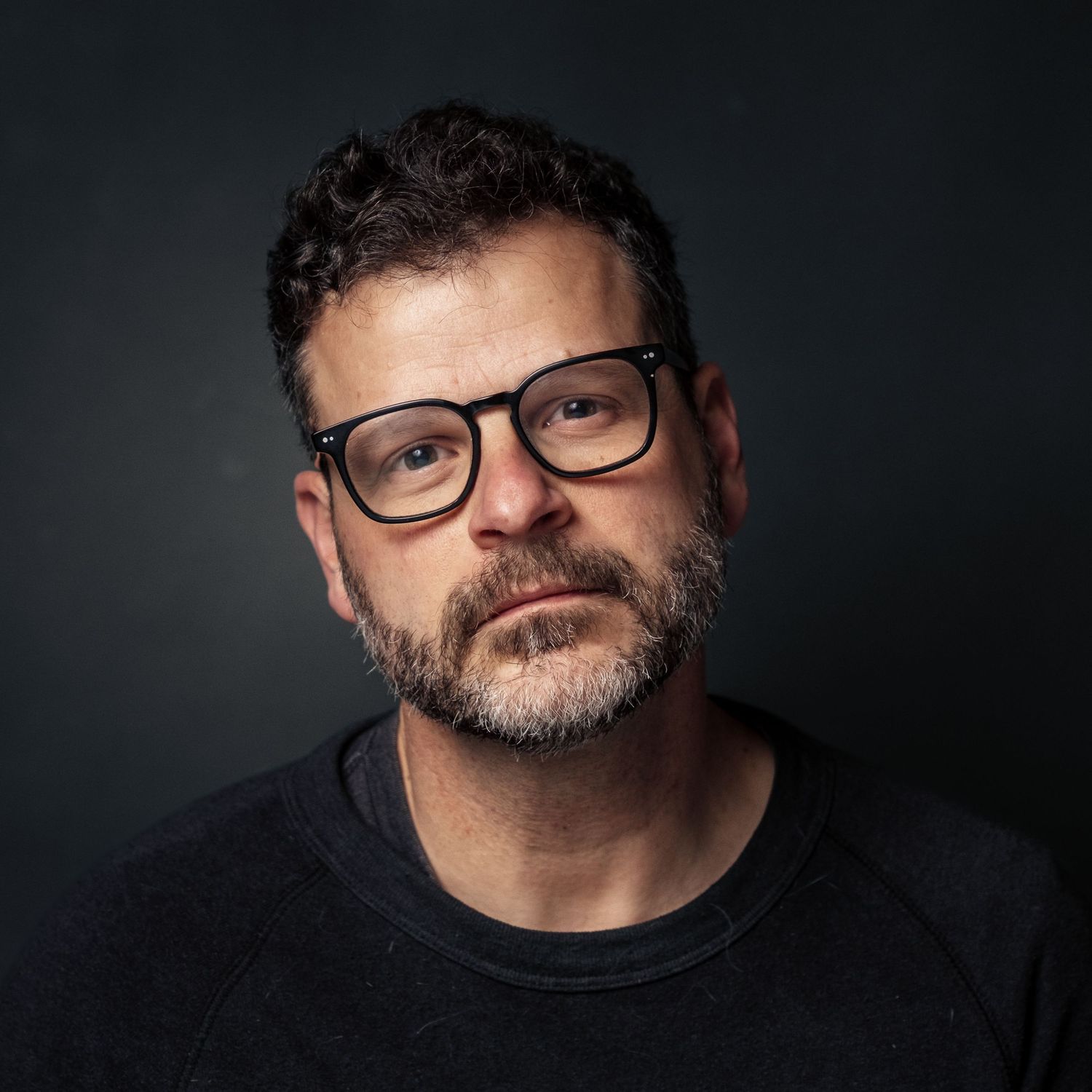 Portrait of bearded person wearing glasses against dark background in dramatic lighting effect.