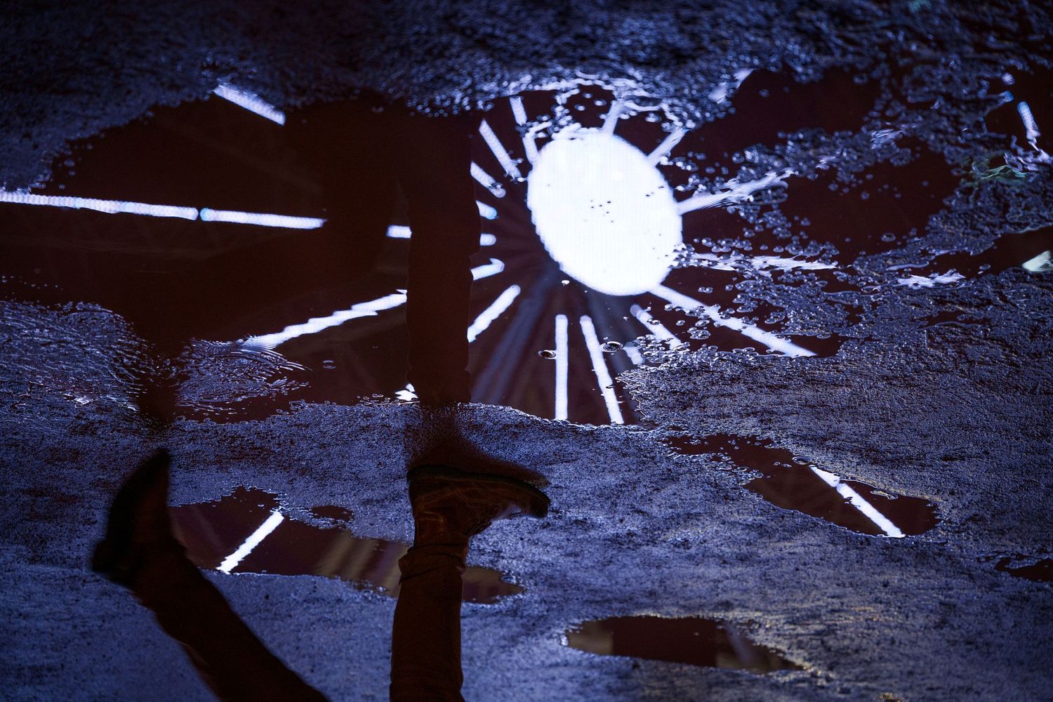 Reflet des lumières d'une grande roue au travers d'une flaque d'eau. L'image est inversée et on voit les jambes d'un passant marcher à côté.