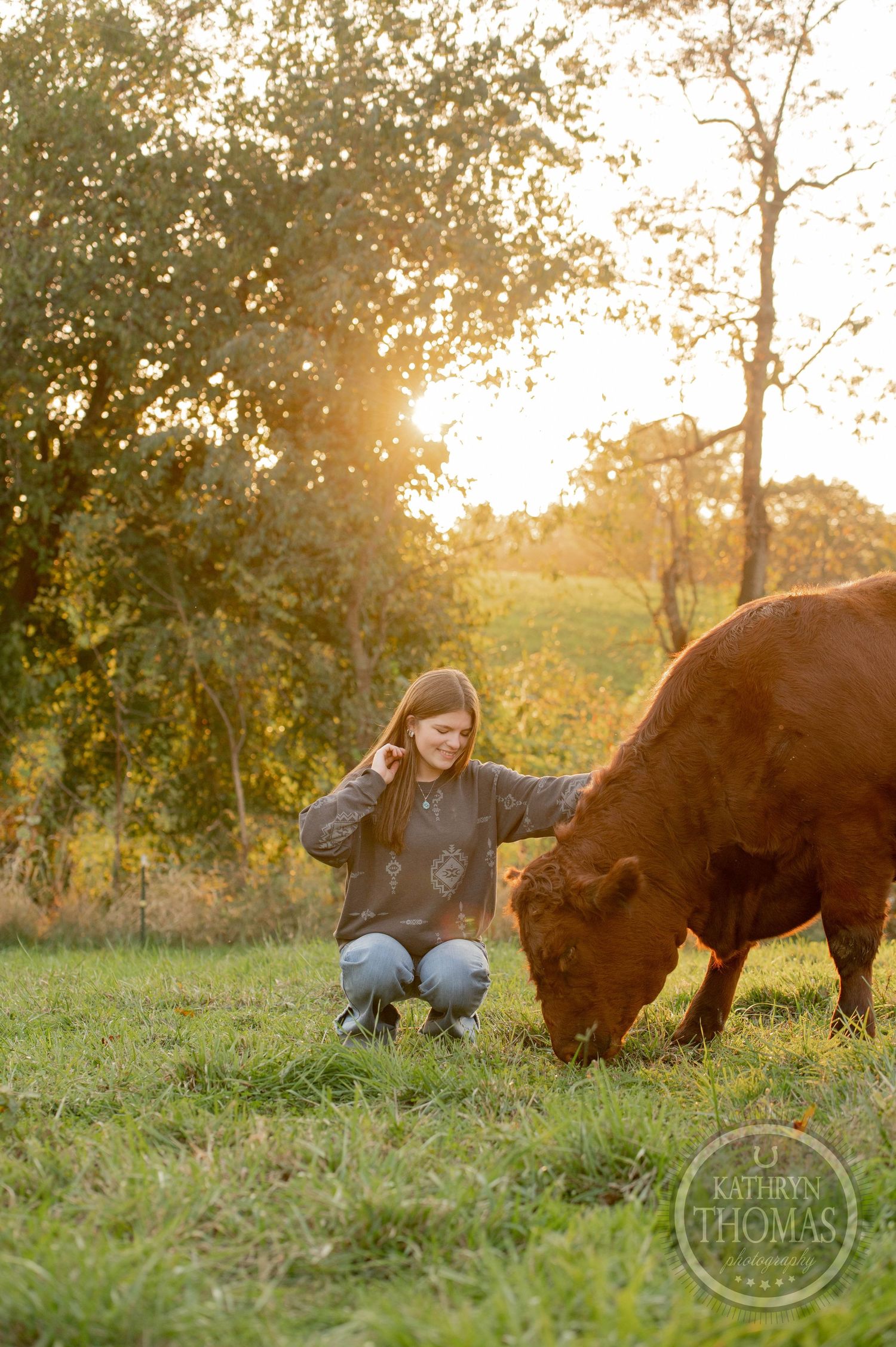 Someone kneels in grass beside a grazing brown cow during golden hour.