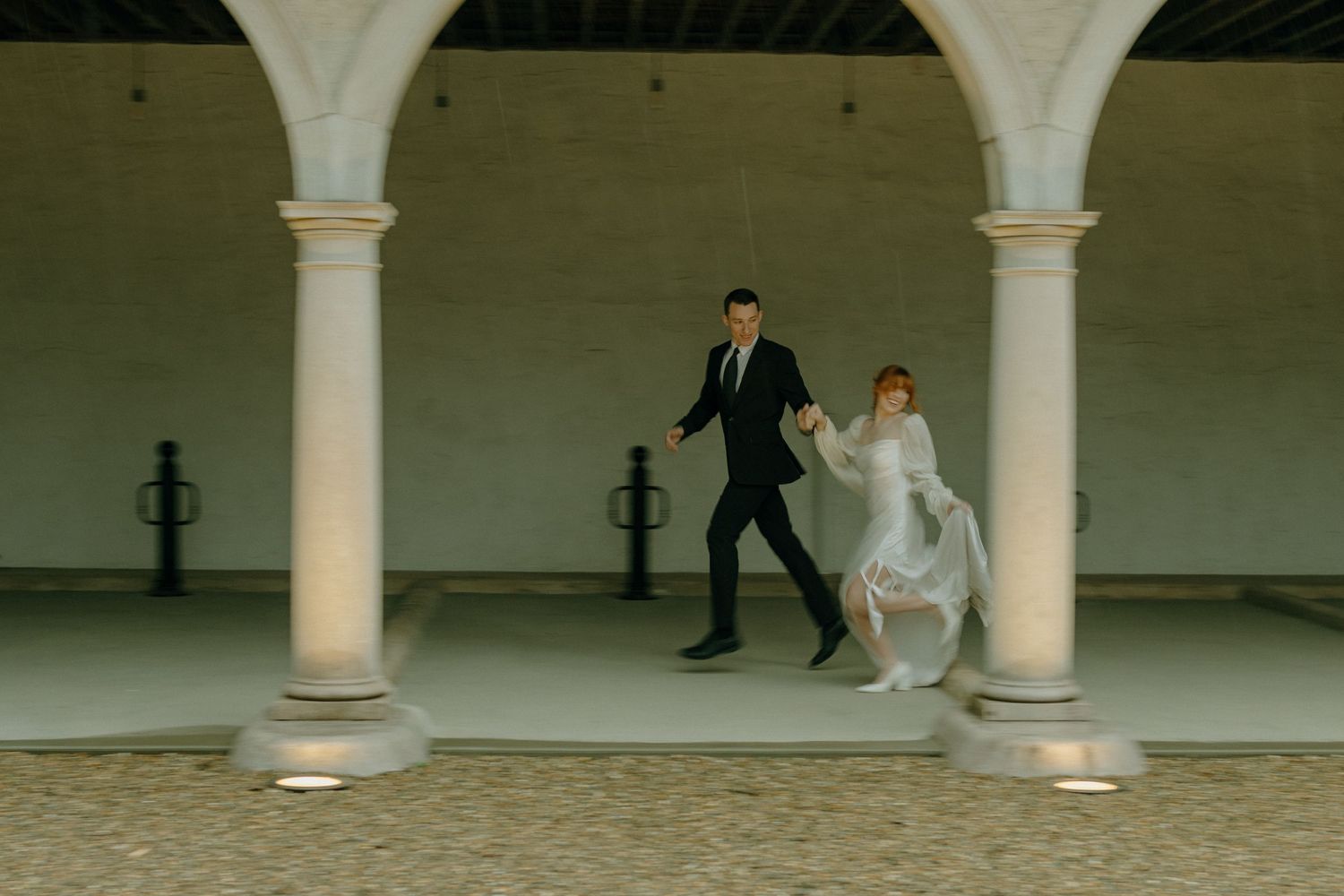 A couple in wedding attire dances between white columns in an outdoor courtyard setting.
