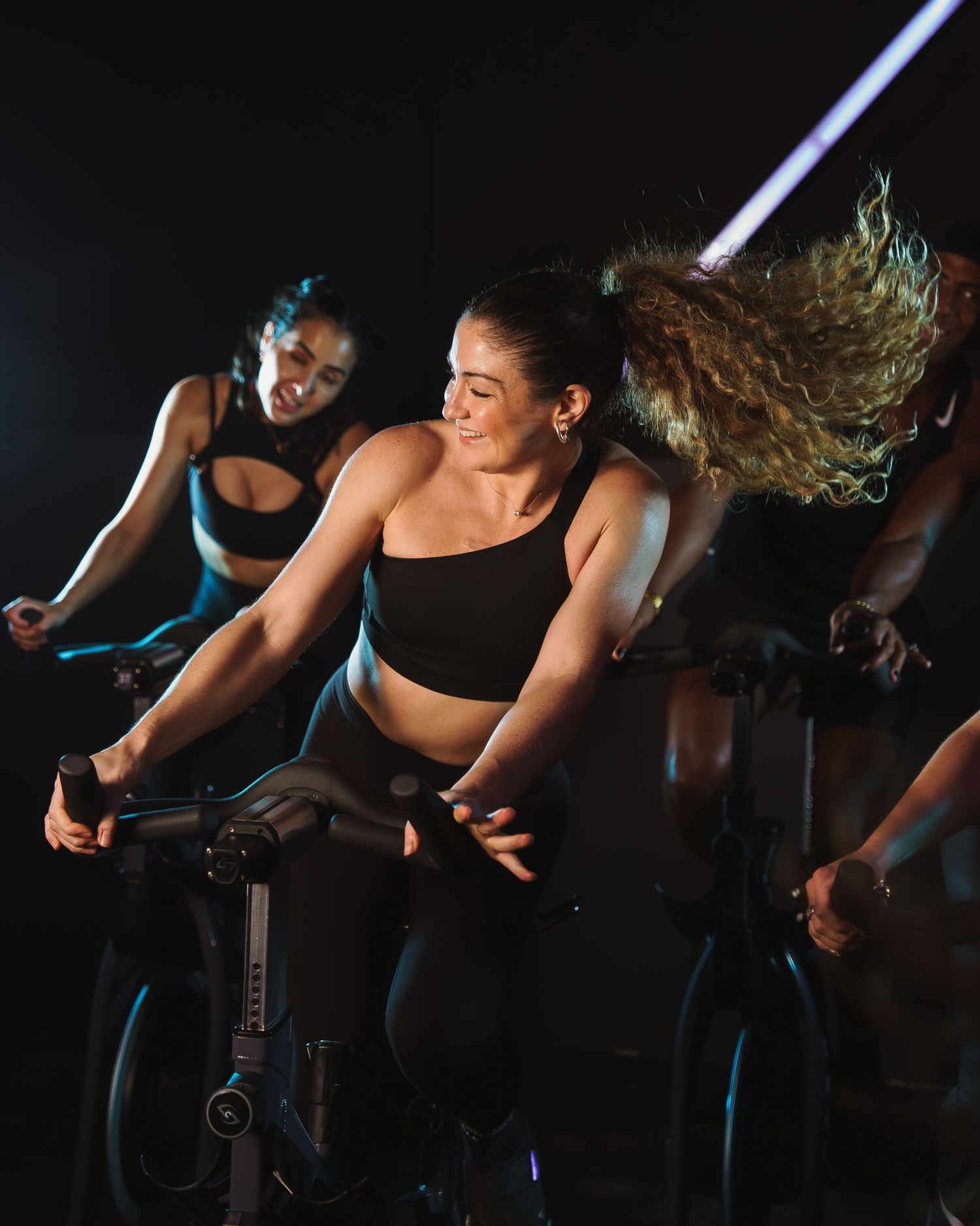Energetic fitness enthusiasts ride indoor cycling bikes during an intense spin class workout in a dark studio.