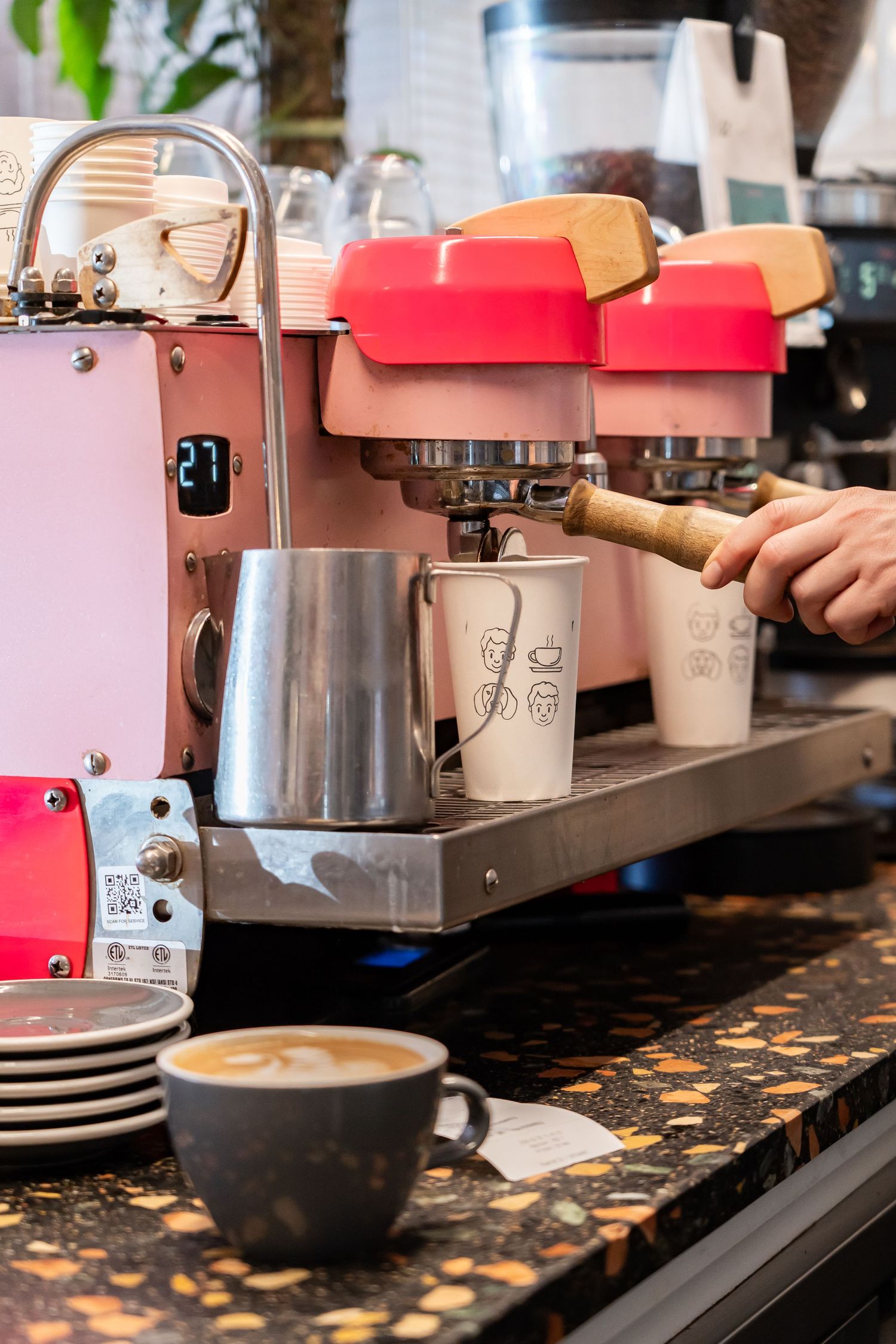 A barista operates a pink espresso machine with red accents while preparing coffee drinks at a cafe counter.
